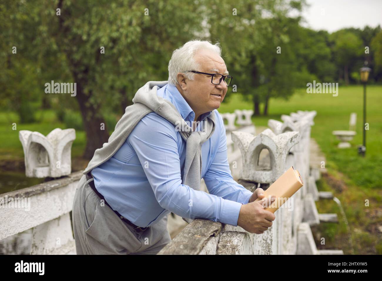 Nachdenklicher älterer Mann, der ein Buch hält, das auf der Brücke im Park steht und nach vorne blickt Stockfoto