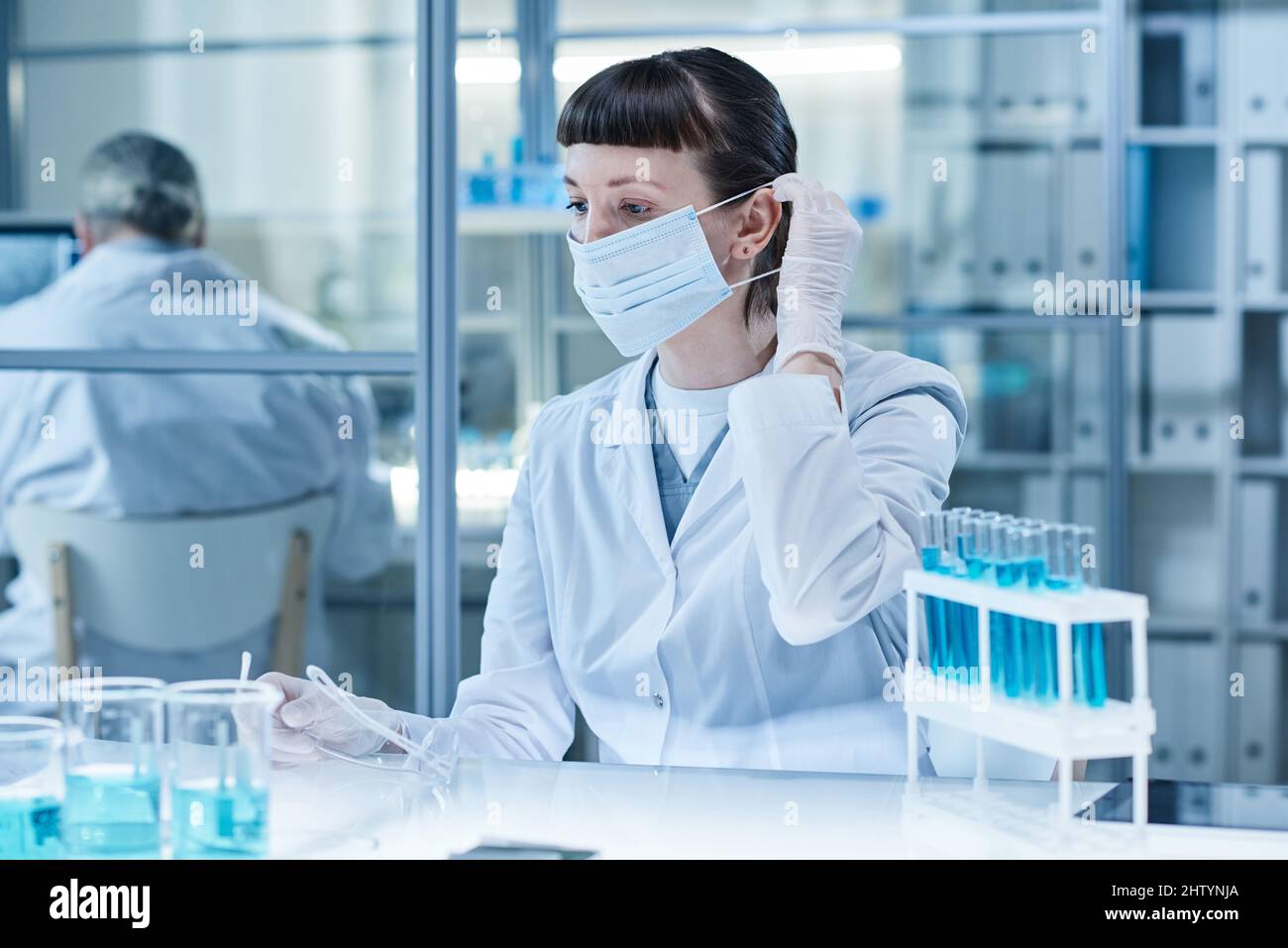 Labortechniker in Maske sitzt im Labor am Tisch und arbeitet mit Proben in Reagenzgläsern Stockfoto