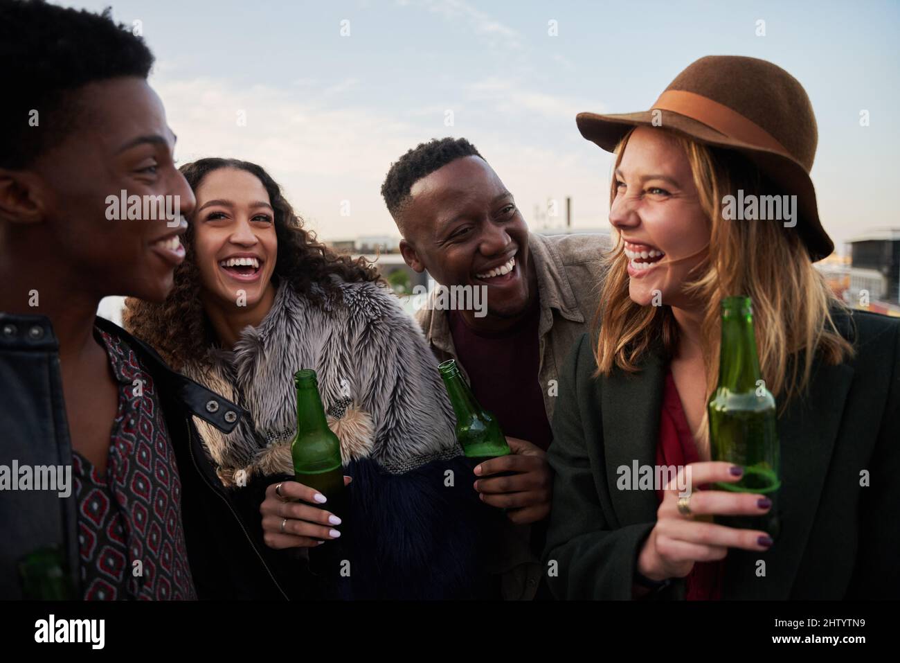 Lachende Gruppe multikultureller junger Erwachsener, die sich auf einer Dachterrasse in der Stadt treffen. Getränke in der Hand, lächelt mit Freunden. Stockfoto