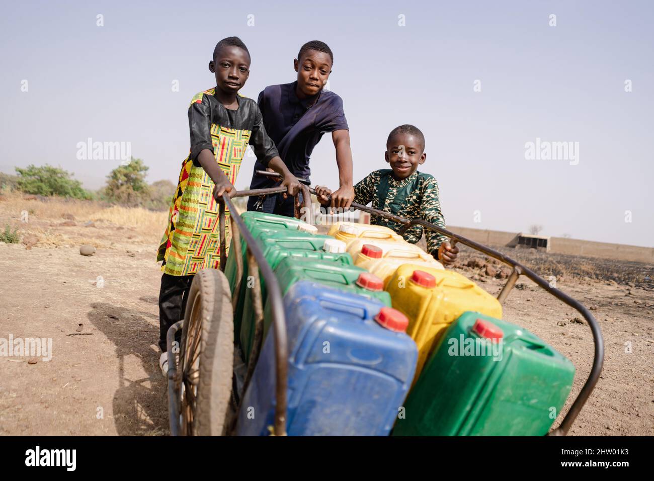 Gruppe von schwarzen Jungen, die schwere Wasserdosen mit einem einfachen Wagen im subsaharischen Afrika tragen; Kinderarbeit Konzept Stockfoto