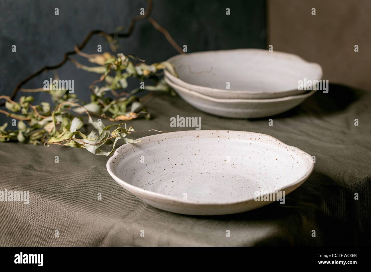 Set aus leerer Keramik weiß gesprenkelt Schüsseln Teller stehen auf dunkelgrünem Tischtuch mit trockenen Ästen Stockfoto