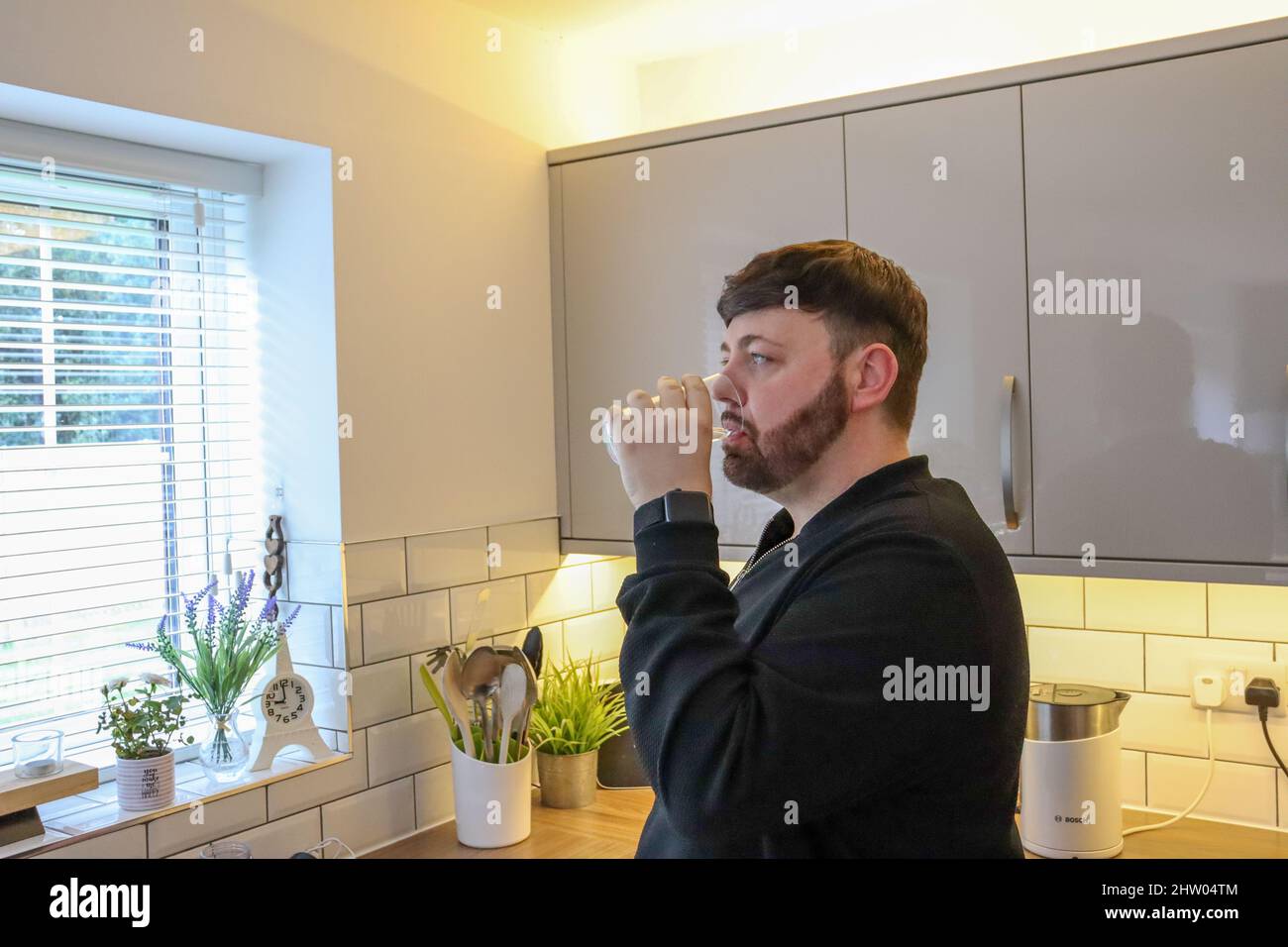 Junger Mann mit fabelhaften Haaren, der in einer modernen Küche ein Glas Wasser trinkt Stockfoto