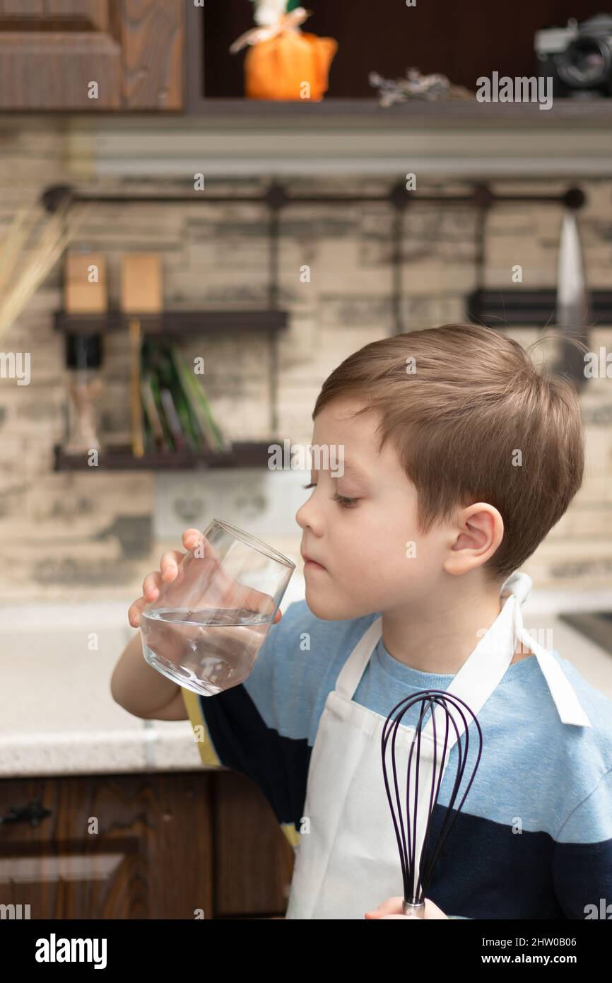 Ein süßer Junge von 7 Jahren in einer Schürze mit einem Schneebesen in der Hand trinkt Wasser aus einem Glas in der Küche vor dem Hintergrund von Küchenutensilien. Se Stockfoto