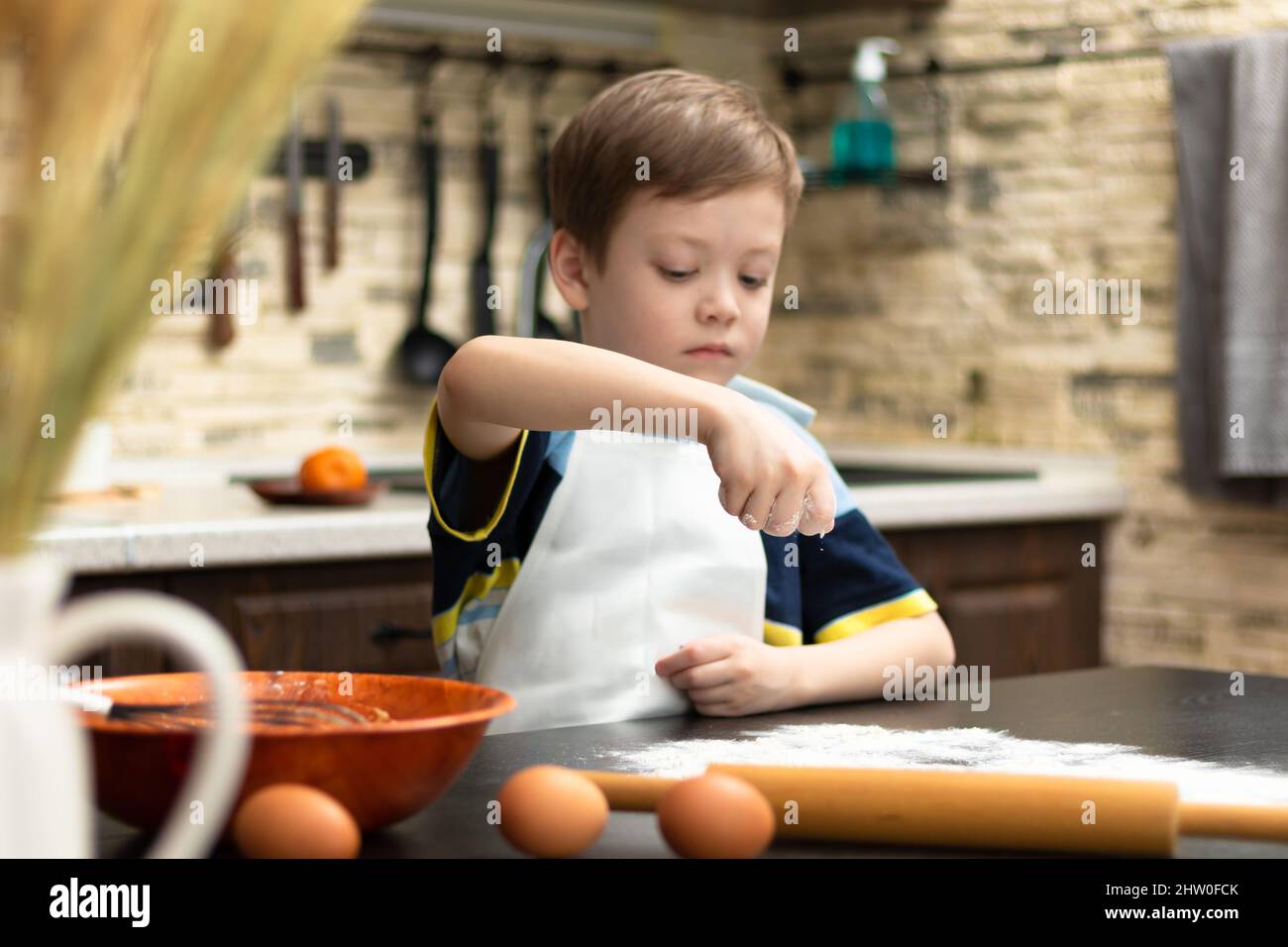 Ein süßer Junge von 7 Jahren in einer Schürze streut Mehl auf den Tisch zu Hause in der Küche vor dem Hintergrund von Küchenutensilien. Selektiver Fokus. Stockfoto