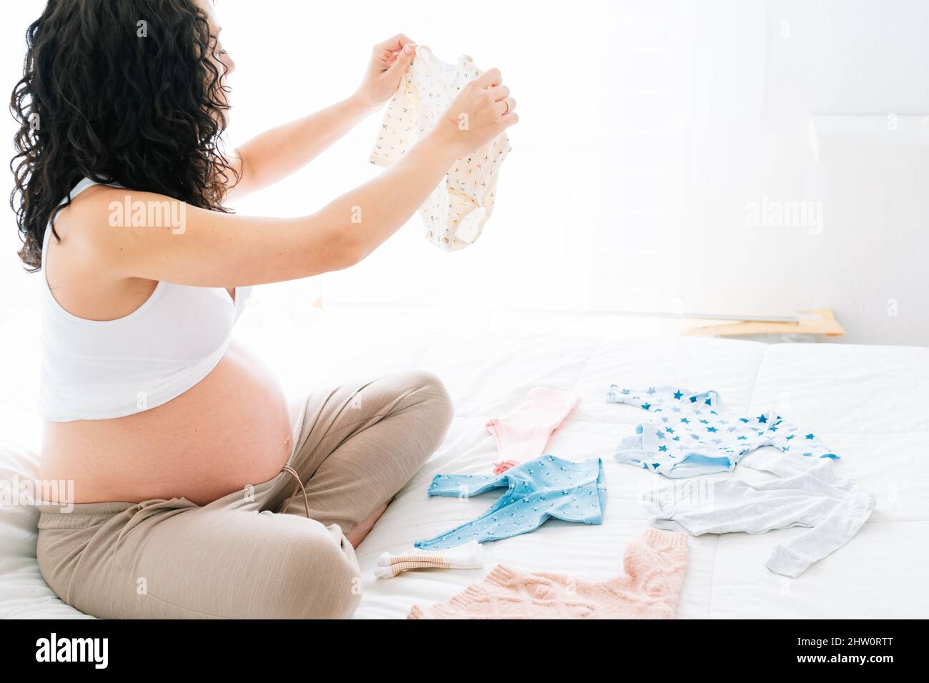 Junge Schwangere, die auf dem Bett sitzt, schaut auf einen der Bodys ihres zukünftigen Babys. Organisation des Zimmers und Vorbereitung der Kleidung der Ne Stockfoto