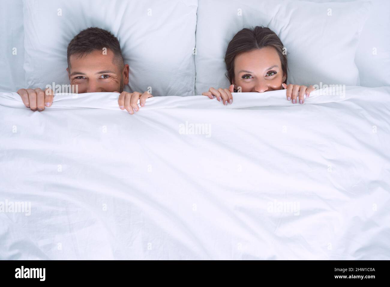 Schönes glückliches Paar im Bett versteckt und bedeckt Gesicht mit weißer Decke. Beziehungskonzept. Speicherplatz kopieren Stockfoto