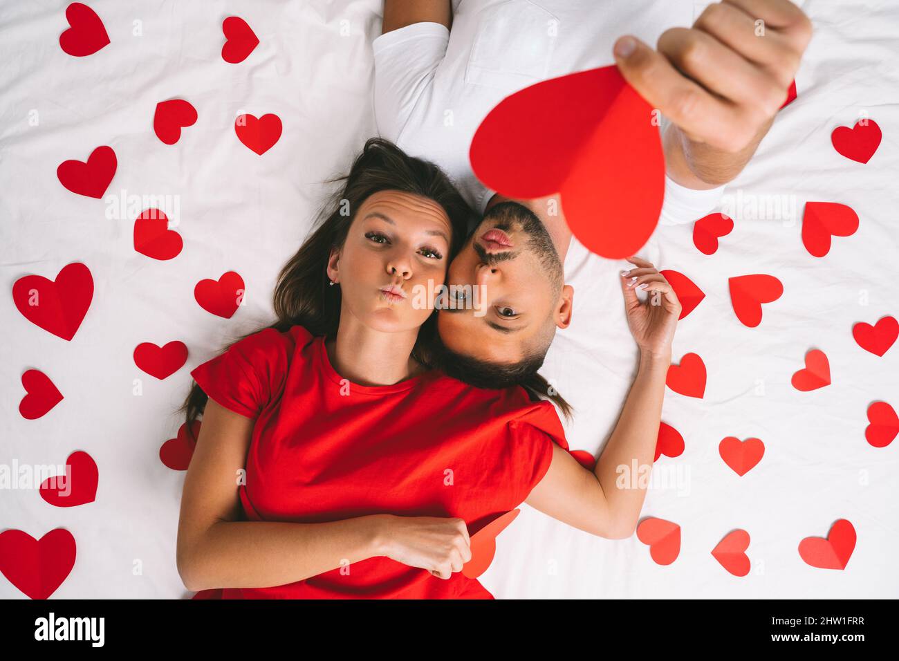 Draufsicht auf ein wunderschönes junges verliebtes Paar, das auf einem weißen Bett mit roten Herzen liegt. Liebe und Beziehungen Stockfoto