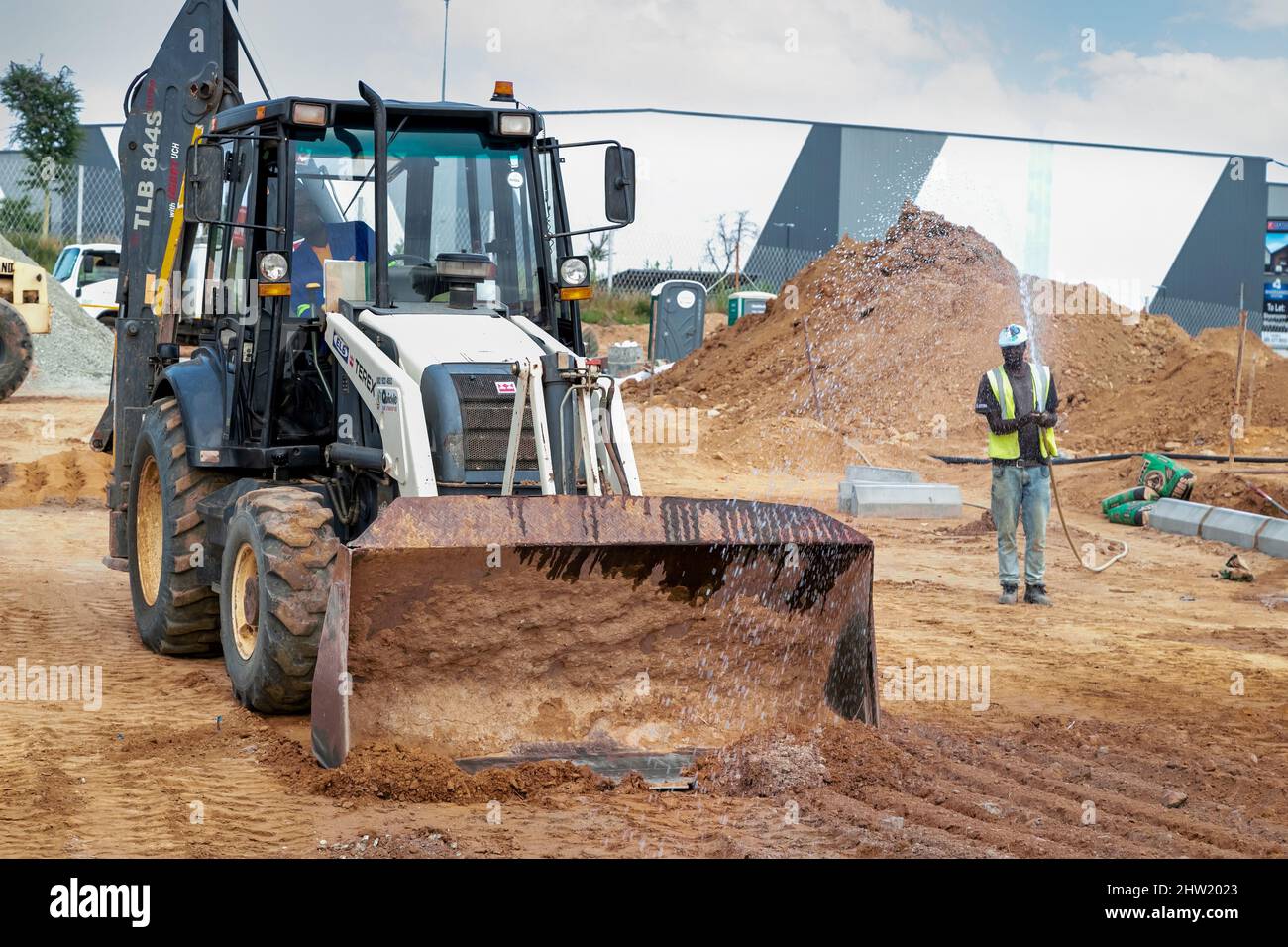 Kapstadt, Südafrika, 24.. Februar - 2022: Mann sprüht Wasser vor dem Bagger, um Staub auf der Baustelle zu halten. Stockfoto