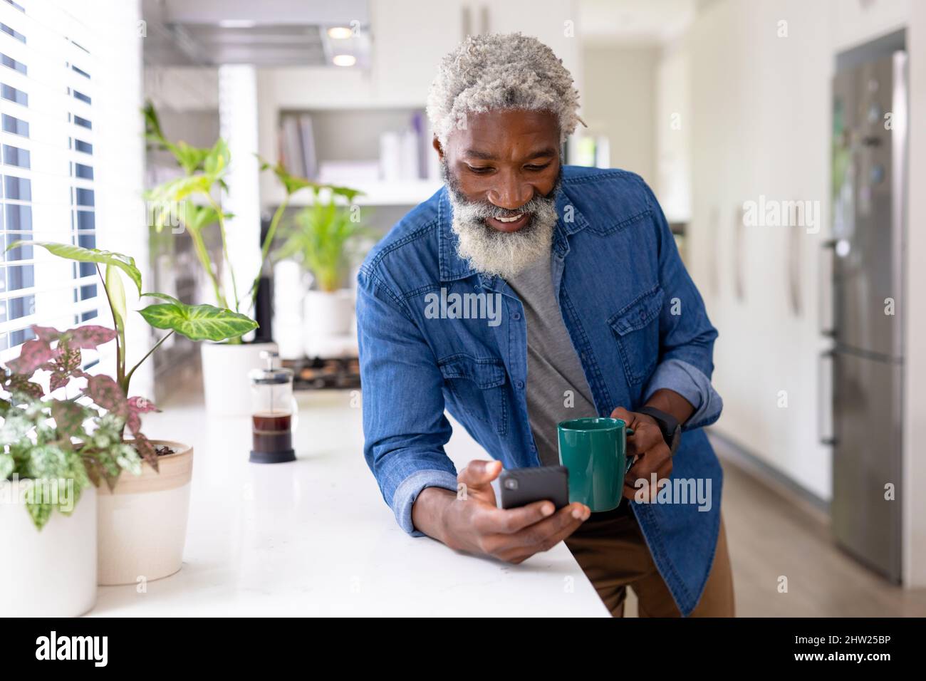 Lächelnder afroamerikanischer Mann, der auf dem Smartphone im Netz surft und den Kaffeebecher auf der Küchentheke hält Stockfoto