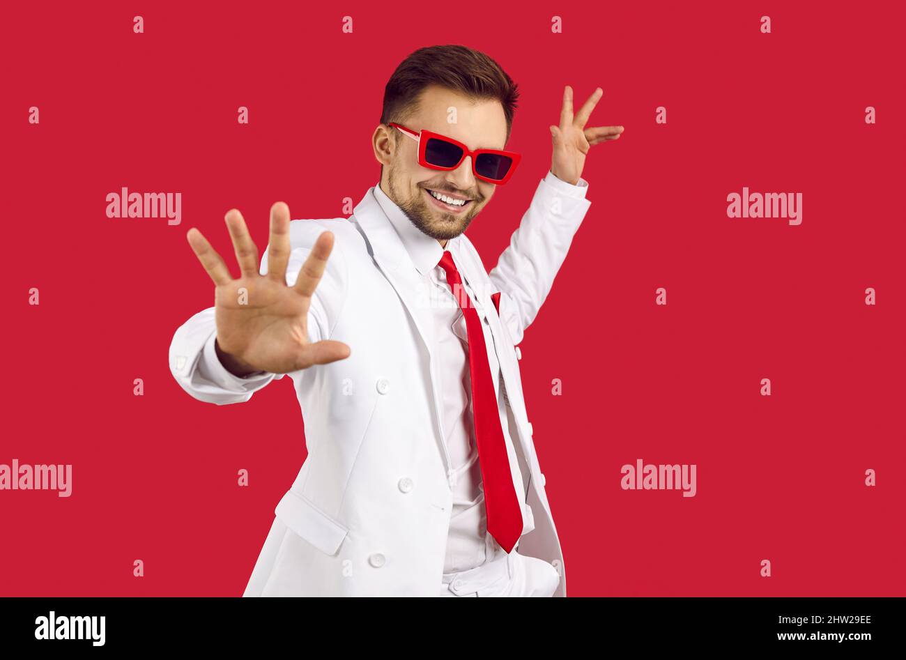 Glücklicher, hübscher Mann mit weißem Anzug und Partybrillen, die auf rotem Hintergrund tanzen Stockfoto