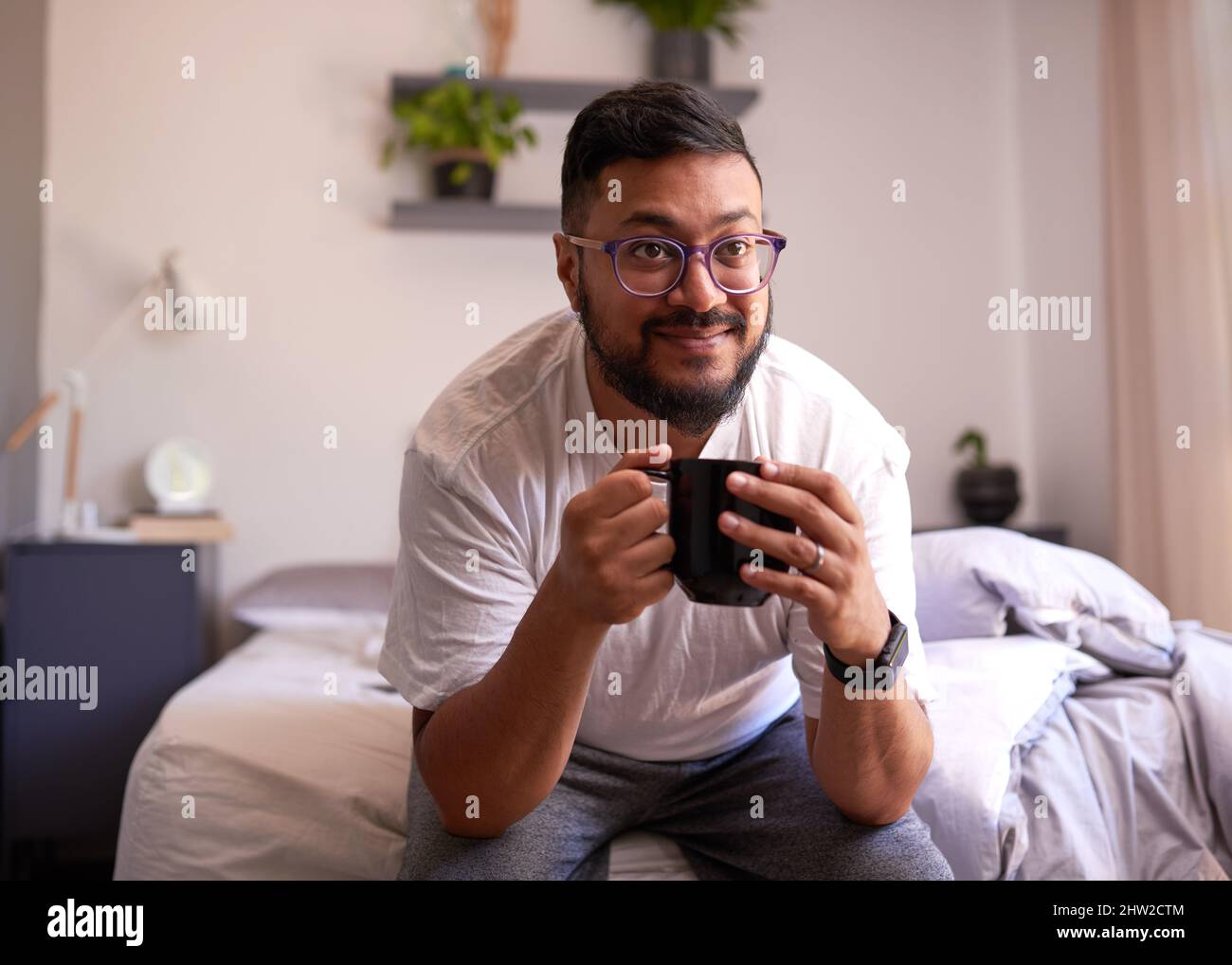Ein junger Erwachsener lächelt am Morgen auf seinem Bett und trinkt Kaffee Stockfoto