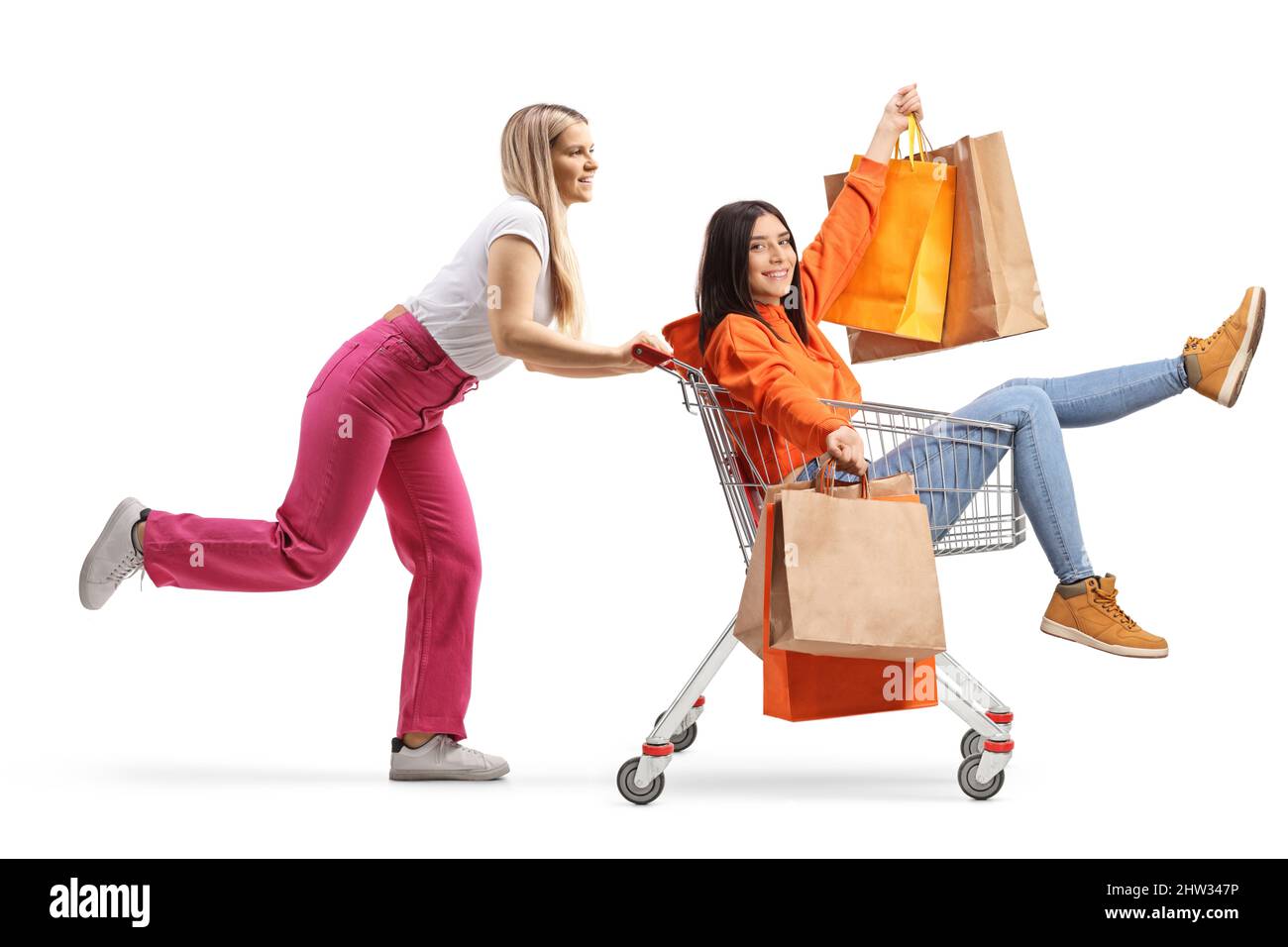 Junge Frau, die ihren Freund in einen Einkaufswagen mit Einkaufstaschen auf weißem Hintergrund isoliert schiebt Stockfoto