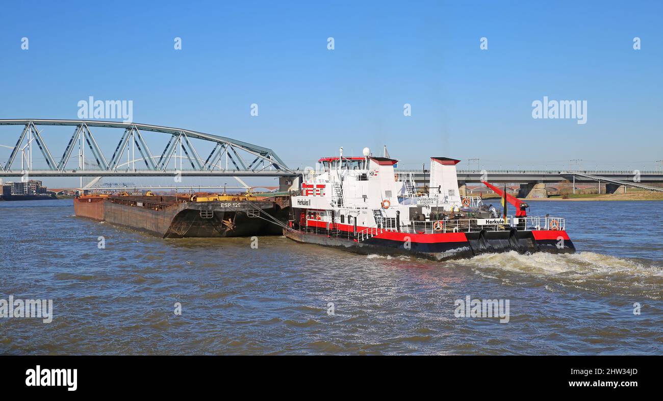 Nijmegen, Niederlande - Februar 27. 2022: Blick auf HGK-Schubschiff herkules-Schiff mit Frachtschiff auf dem Fluss waal gegen blauen Himmel und Eisenbahnstahl b Stockfoto