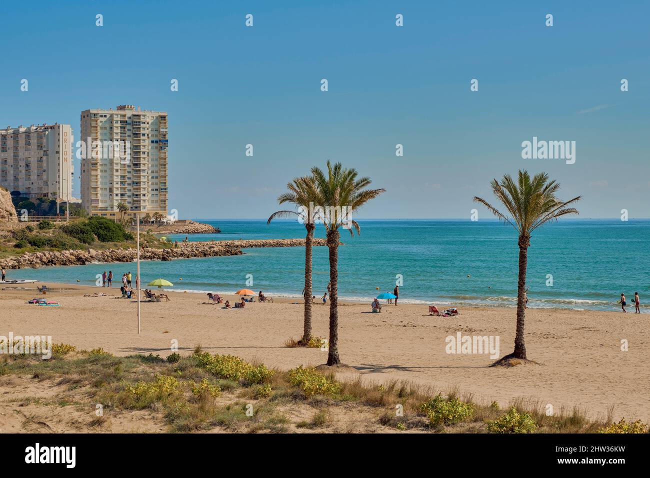Menschen, die mit ihren Sonnenschirmen auf dem Sand sonnenbaden und am Strand von Racó in der Küstenstadt Cullera in der Provinz Valencia, Spanien, Europehor baden Stockfoto