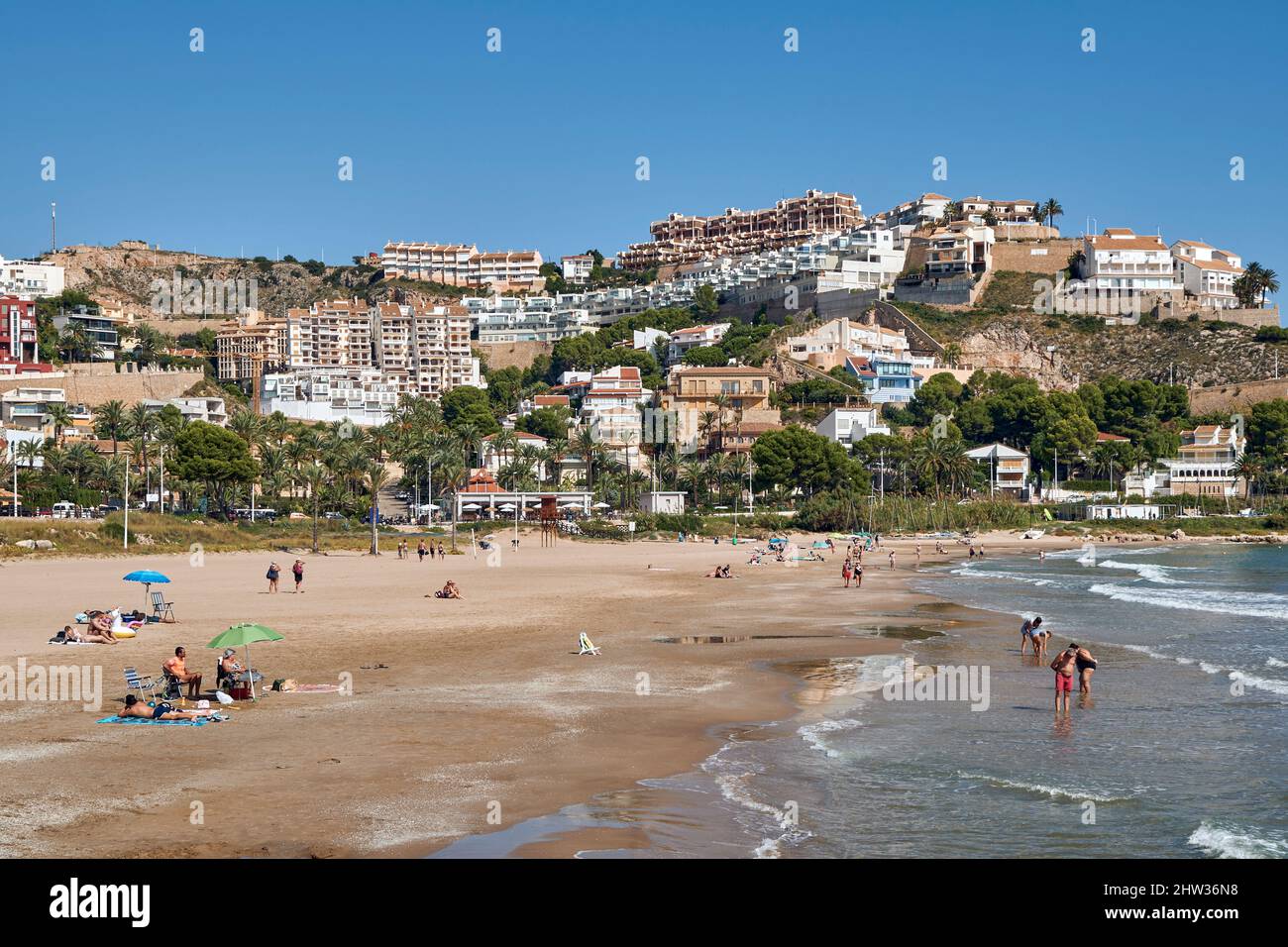 Menschen, die sich mit ihren Sonnenschirmen auf dem Sand sonnen und in der Platja de l'Illa der Küstenstadt Cullera in der Provinz Valencia, Spanien, baden, Stockfoto