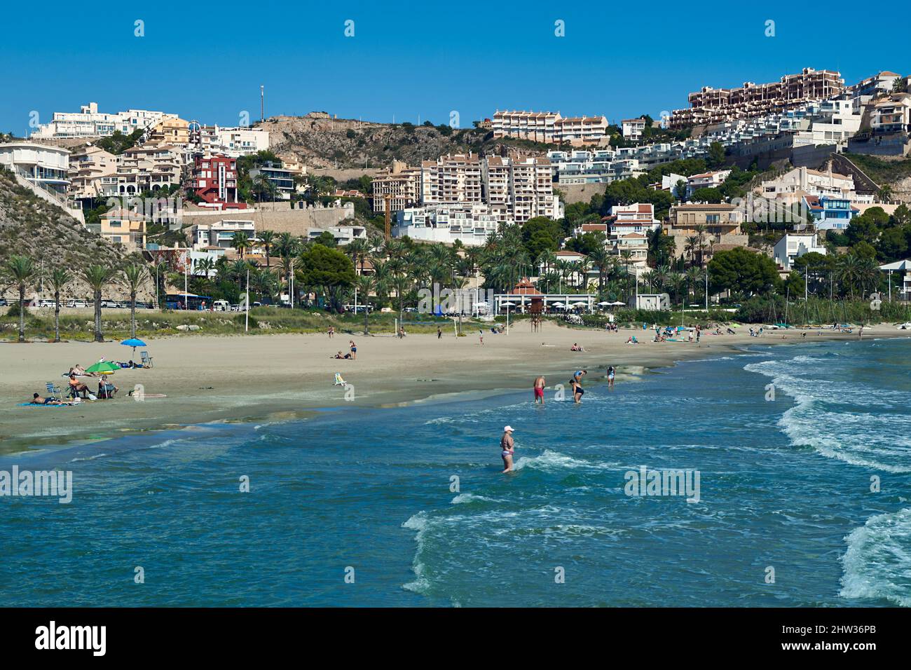 Menschen, die sich mit ihren Sonnenschirmen auf dem Sand sonnen und in der Platja de l'Illa der Küstenstadt Cullera in der Provinz Valencia, Spanien, baden, Stockfoto