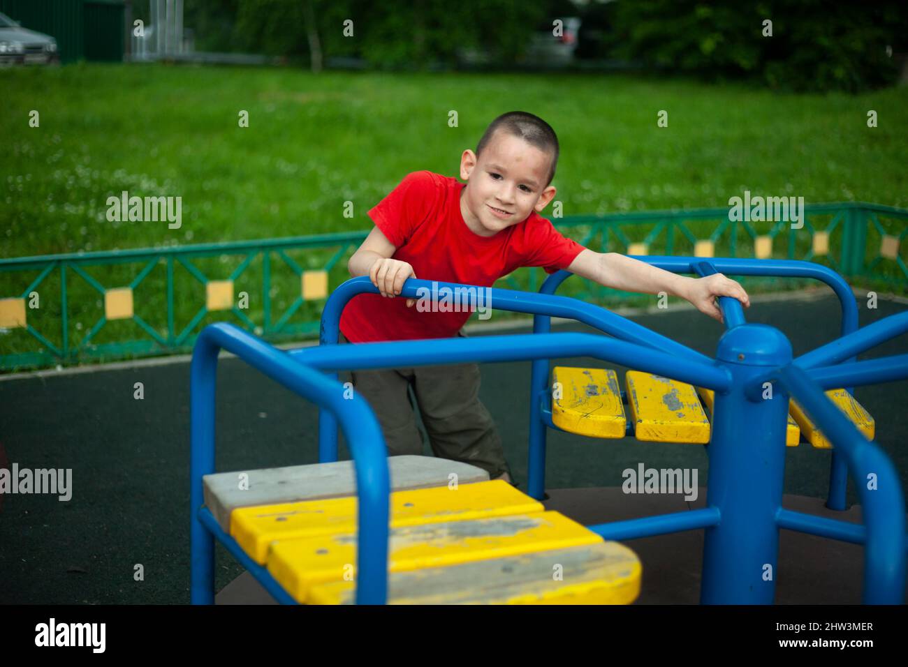 Kind im Karussell. Junge dreht sich auf dem Karussell für Kinder auf dem Spielplatz. Junior Schuljunge spielt allein. Kind in rotem T-Shirt mit kurzem Haarschnitt. Mann Stockfoto