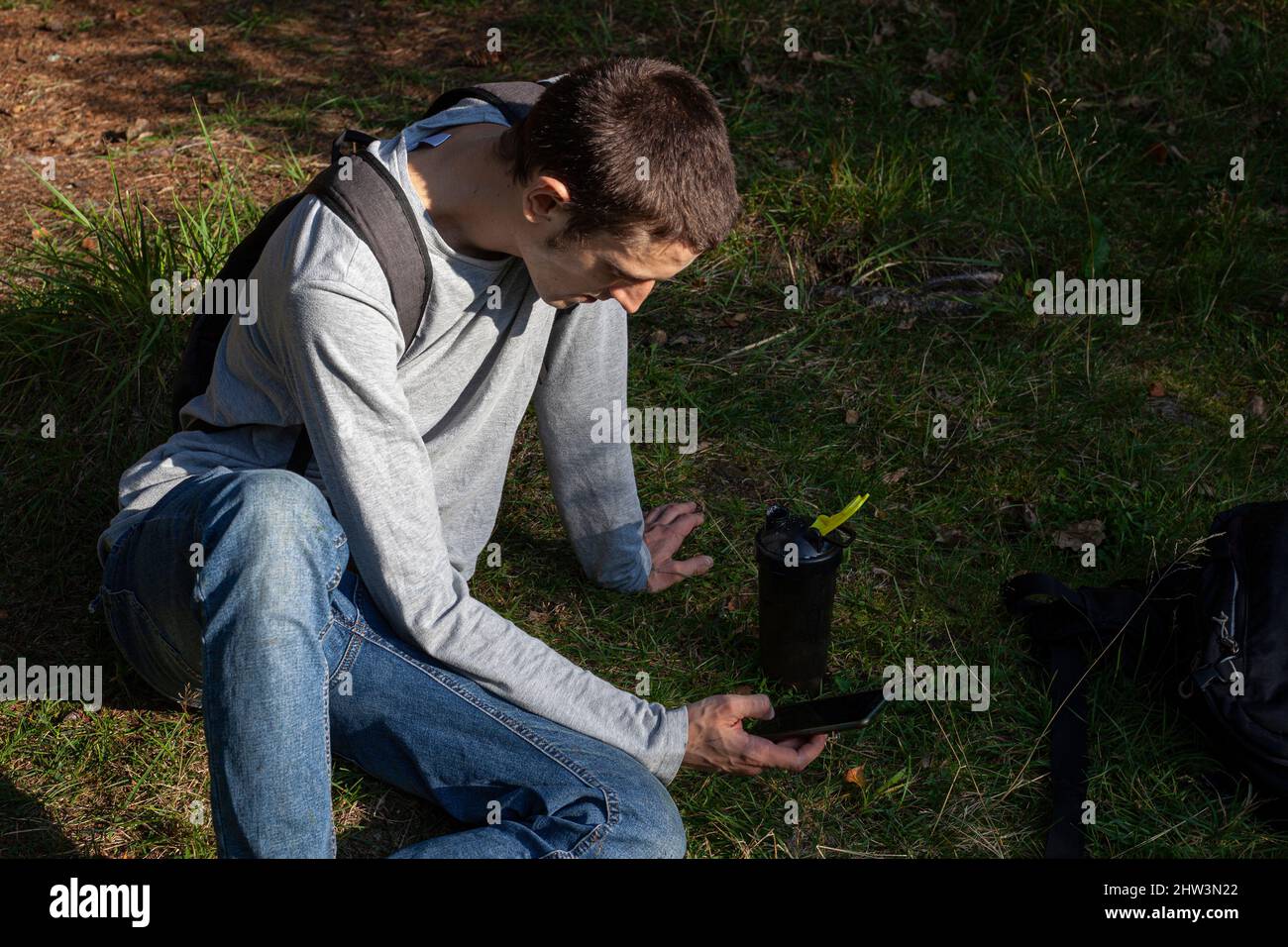 Der Kerl ruht sich im Park aus und schaut auf das Smartphone. Ein junger Mann entspannt sich im Wald. Eine interessante Person mit europäischem Auftritt. Camping Stockfoto