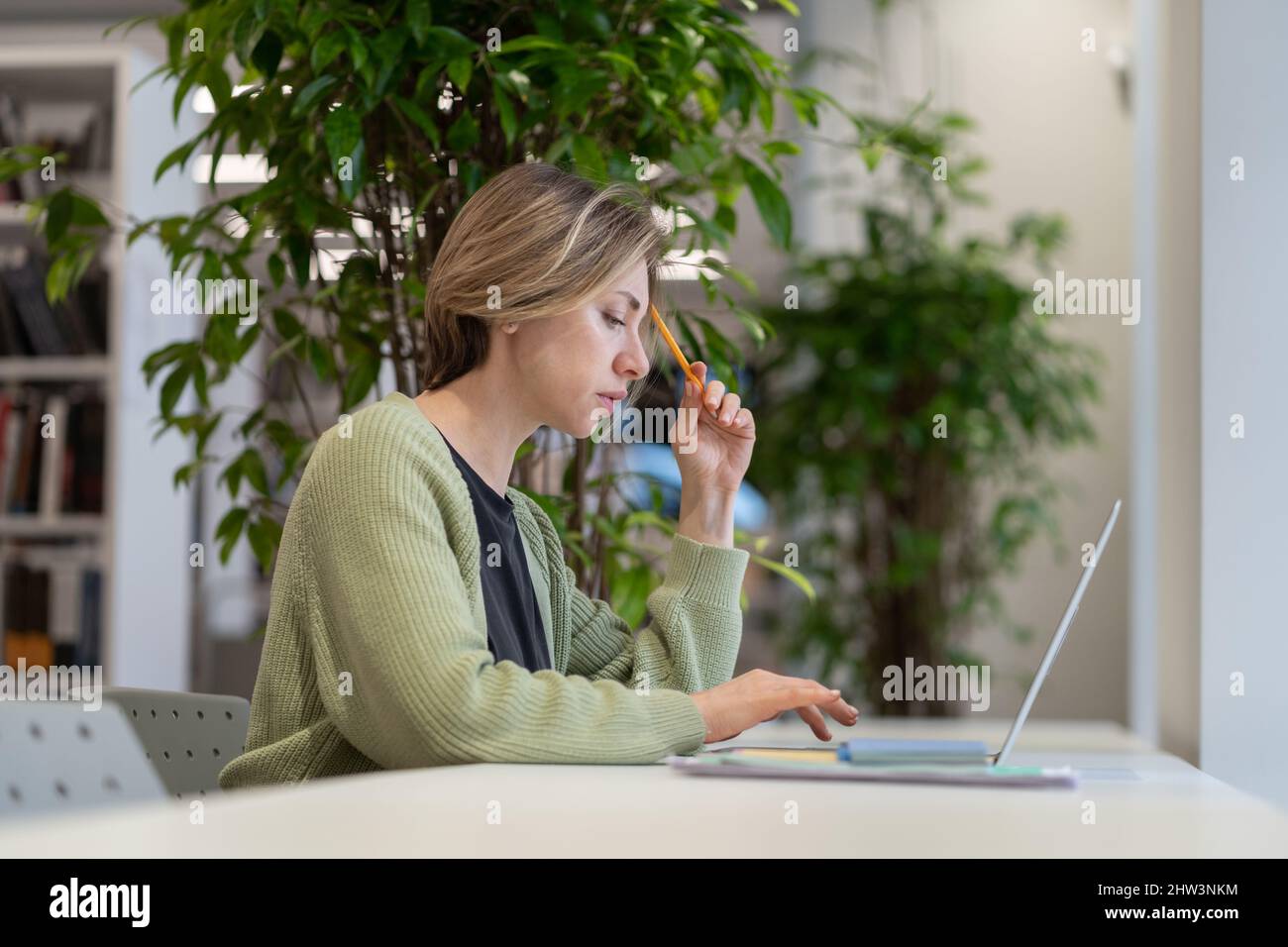 Nachdenkliche Professorin mittleren Alters, die in der Universitätsbibliothek Vorlesungsmaterial auf einem Laptop vorbereitet Stockfoto