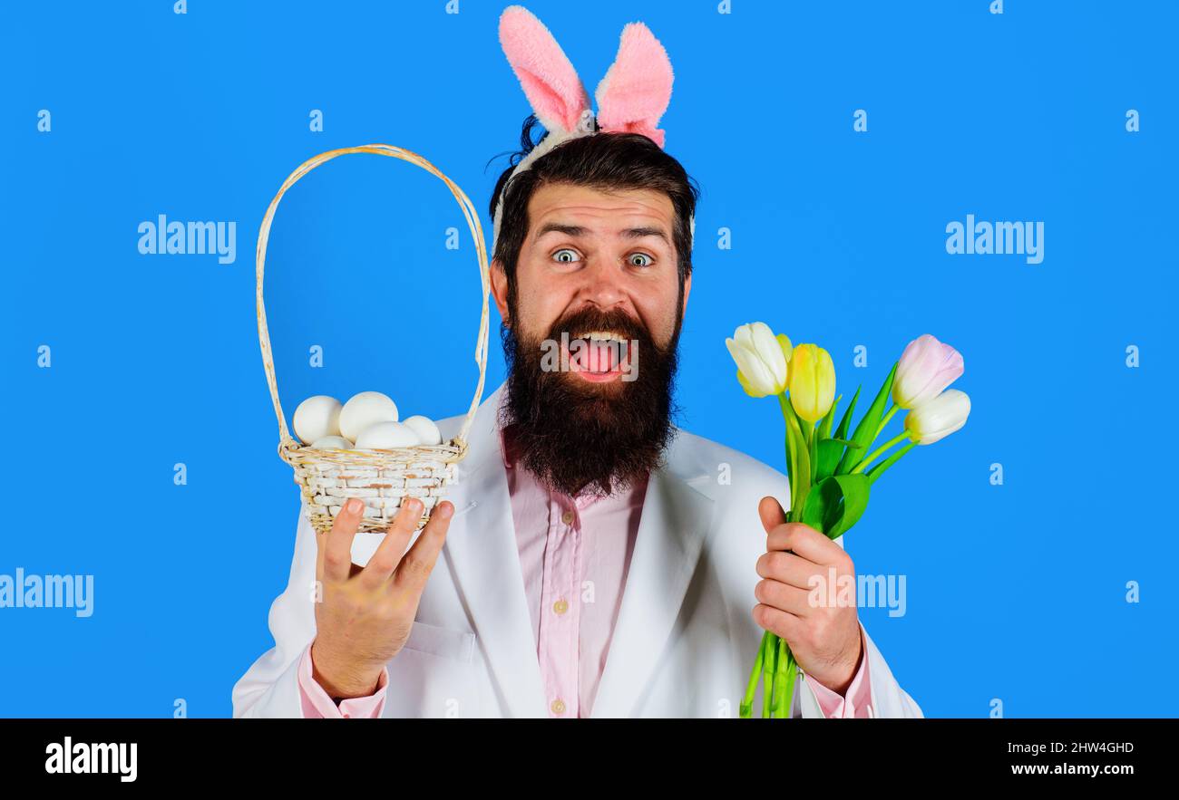Ostertag. Kaninchenmann in Hasenohren mit Blume. Bärtiger Rüde in Anzug hält Korb mit Eiern. Stockfoto