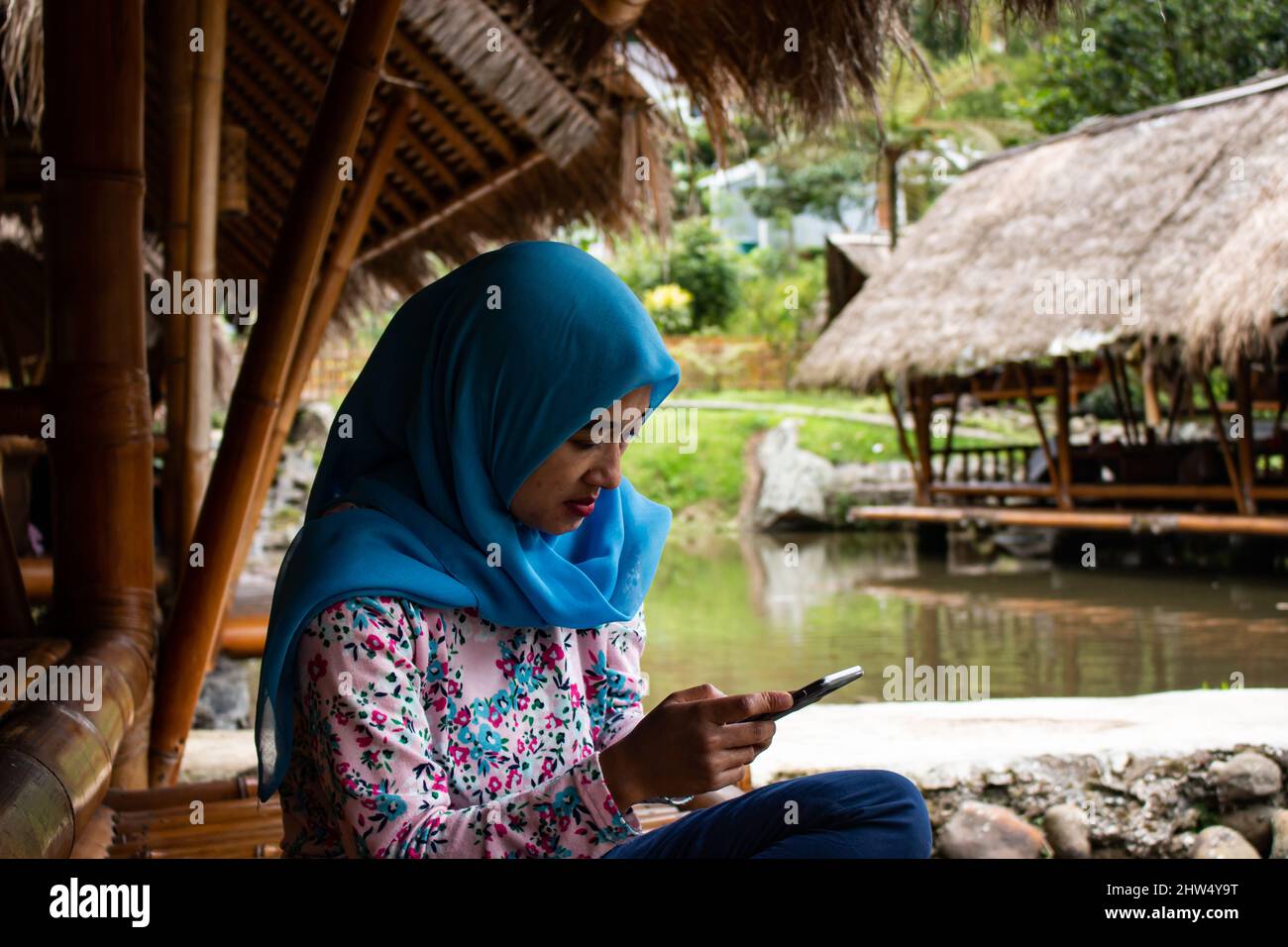 Junge asiatische Frau sitzt auf Bambus im Essbereich Surfen im Internet auf modernen mobilen Gadget, glücklich Millennial Indonesian Mädchen entspannende Textnachricht Stockfoto