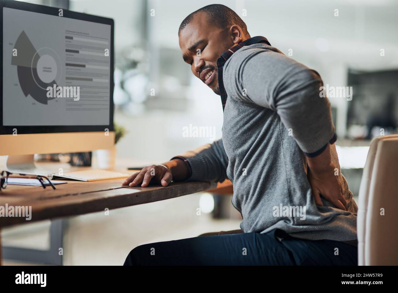 Zu viele Stunden im heißen Sitz. Aufnahme eines jungen Geschäftsmannes, der an Rückenschmerzen leidet, während er an seinem Schreibtisch in seinem Büro arbeitet. Stockfoto