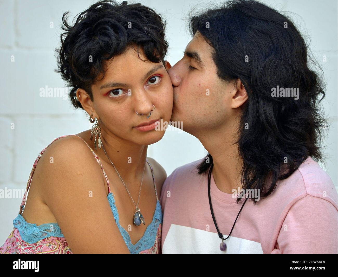 Ein hübscher junger Latino-Mann mit langen Haaren küsst eine schöne junge Latina-Frau mit kurzen Haaren auf der Wange. Stockfoto