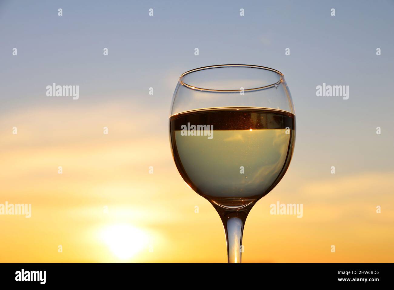 Glas mit Weißwein auf Sonnenuntergangshintergrund, Abendsonne und Himmel mit Wolken spiegeln sich im Glas. Konzept der Feier, Weinindustrie Stockfoto