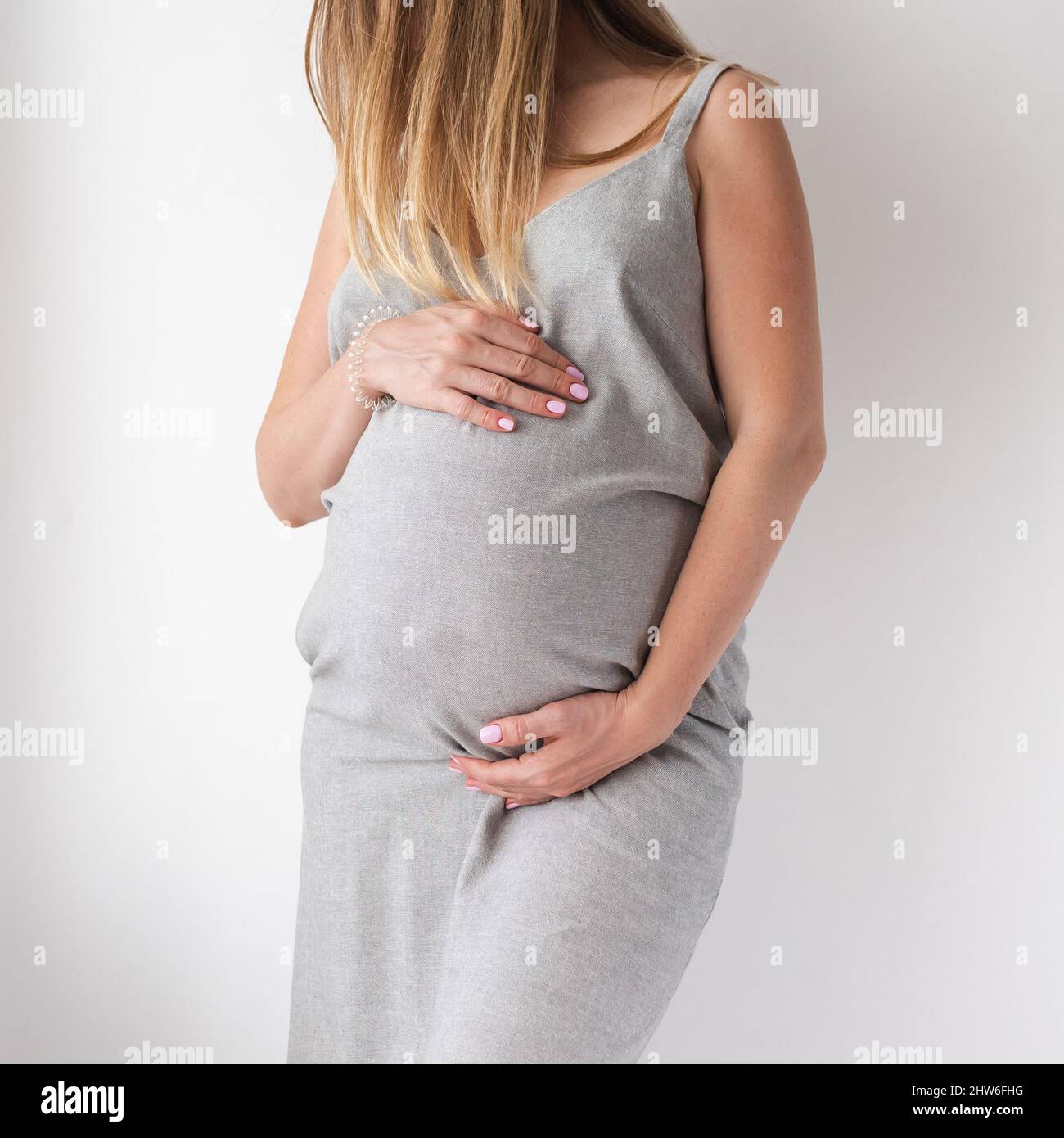 Eine anspruchsvolle Schwangere in einem grauen Kleid steht auf weißem Hintergrund mit den Händen auf dem Bauch, dem Konzept der Weiblichkeit Stockfoto