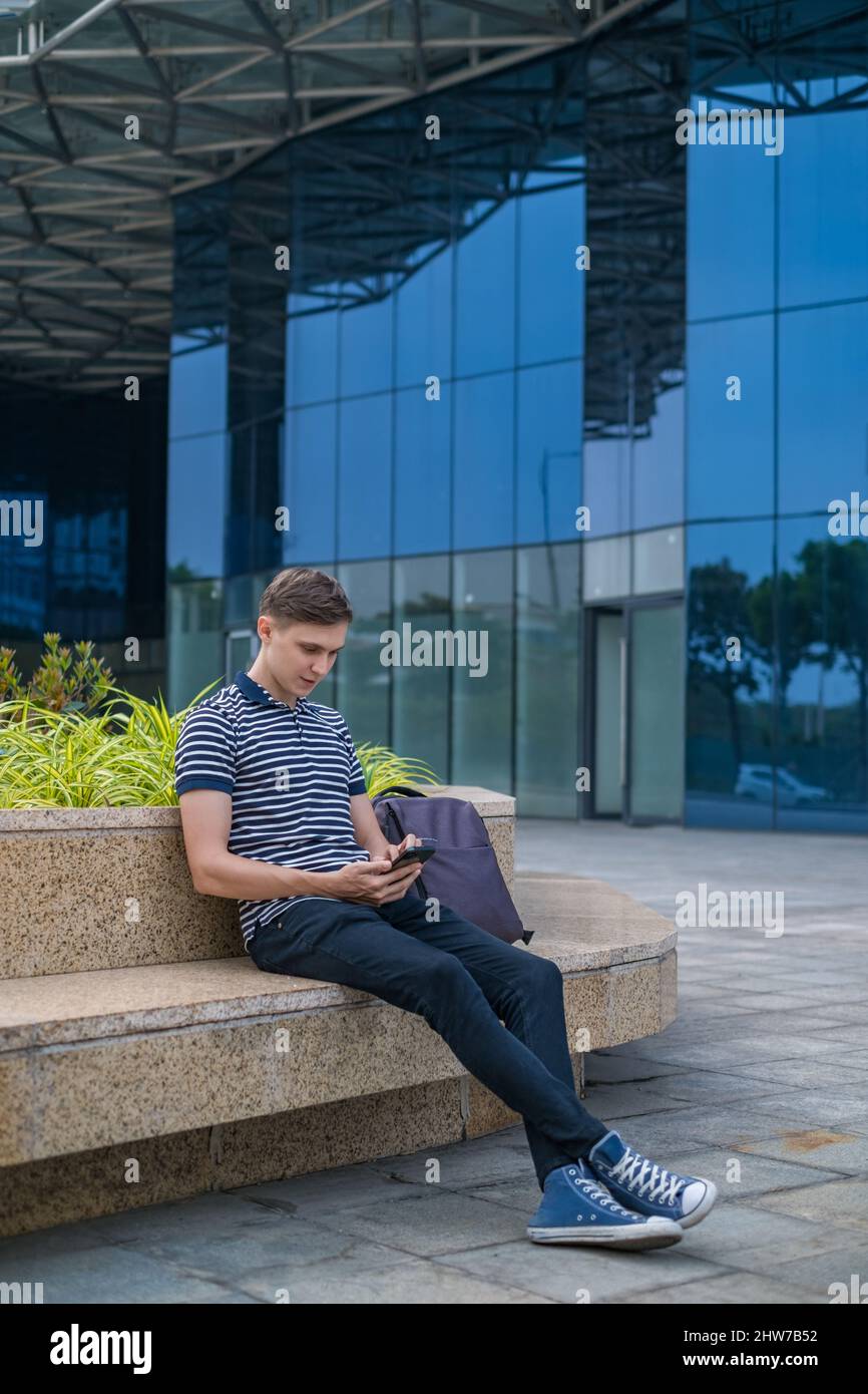 Junger kaukasischer Mann in legerer Kleidung sitzt in der Nähe des modernen Bürogebäudes und schaut auf sein Telefon. Hochwertige Fotos Stockfoto