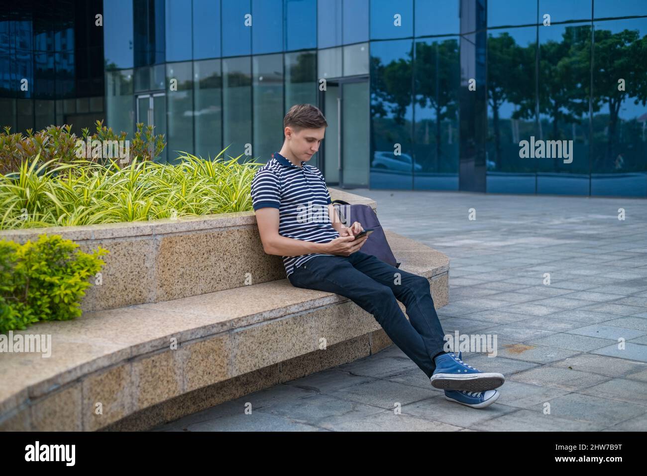 Junger kaukasischer Mann in legerer Kleidung sitzt in der Nähe des modernen Bürogebäudes und schaut auf sein Telefon. Hochwertige Fotos Stockfoto