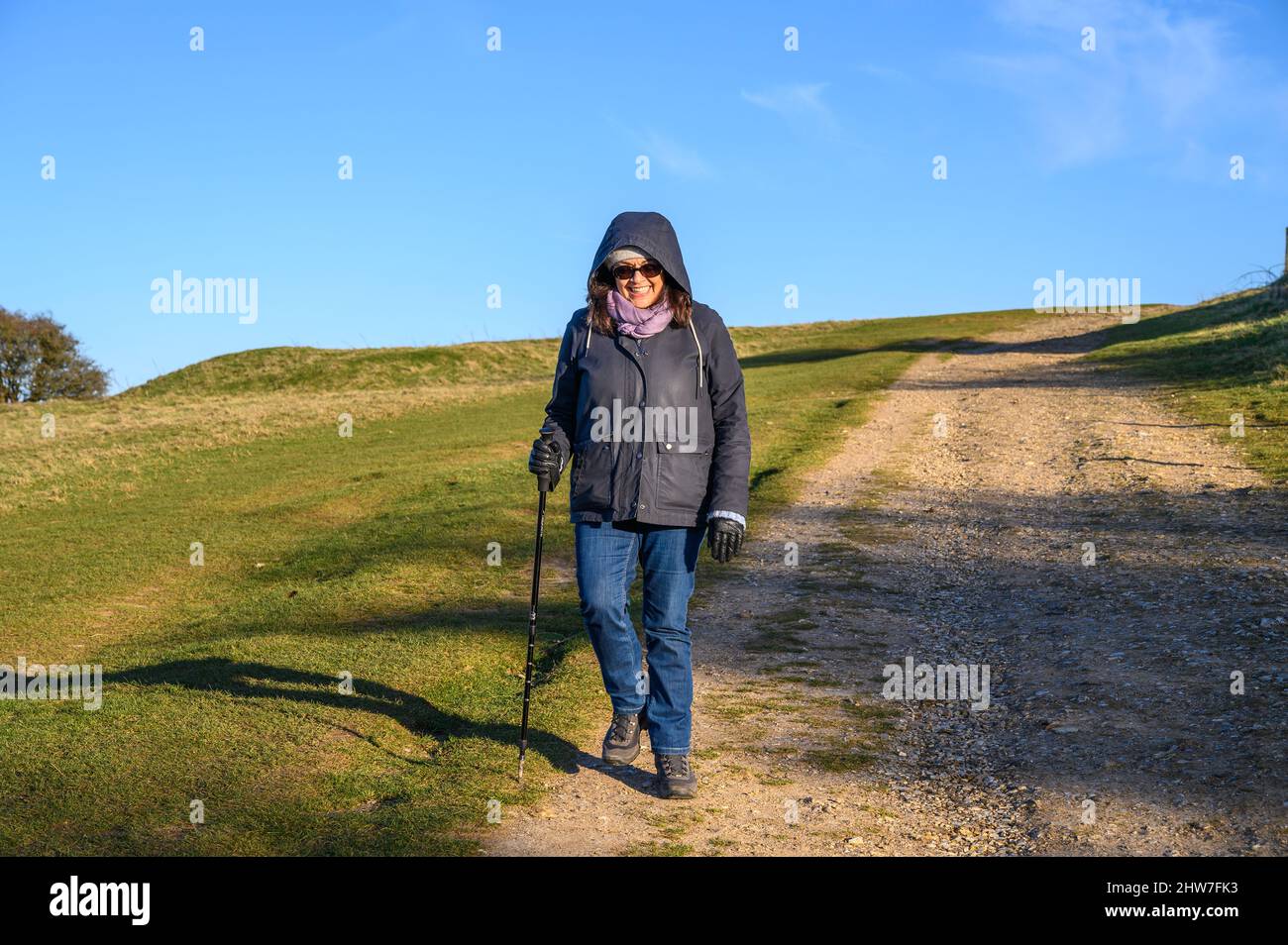 Eine Frau mittleren Alters mit indischer Ethnie, die in niedriger Wintersonne westlich von Ditchling Beacon in East Sussex, England, den South Downs Way entlang geht. Stockfoto