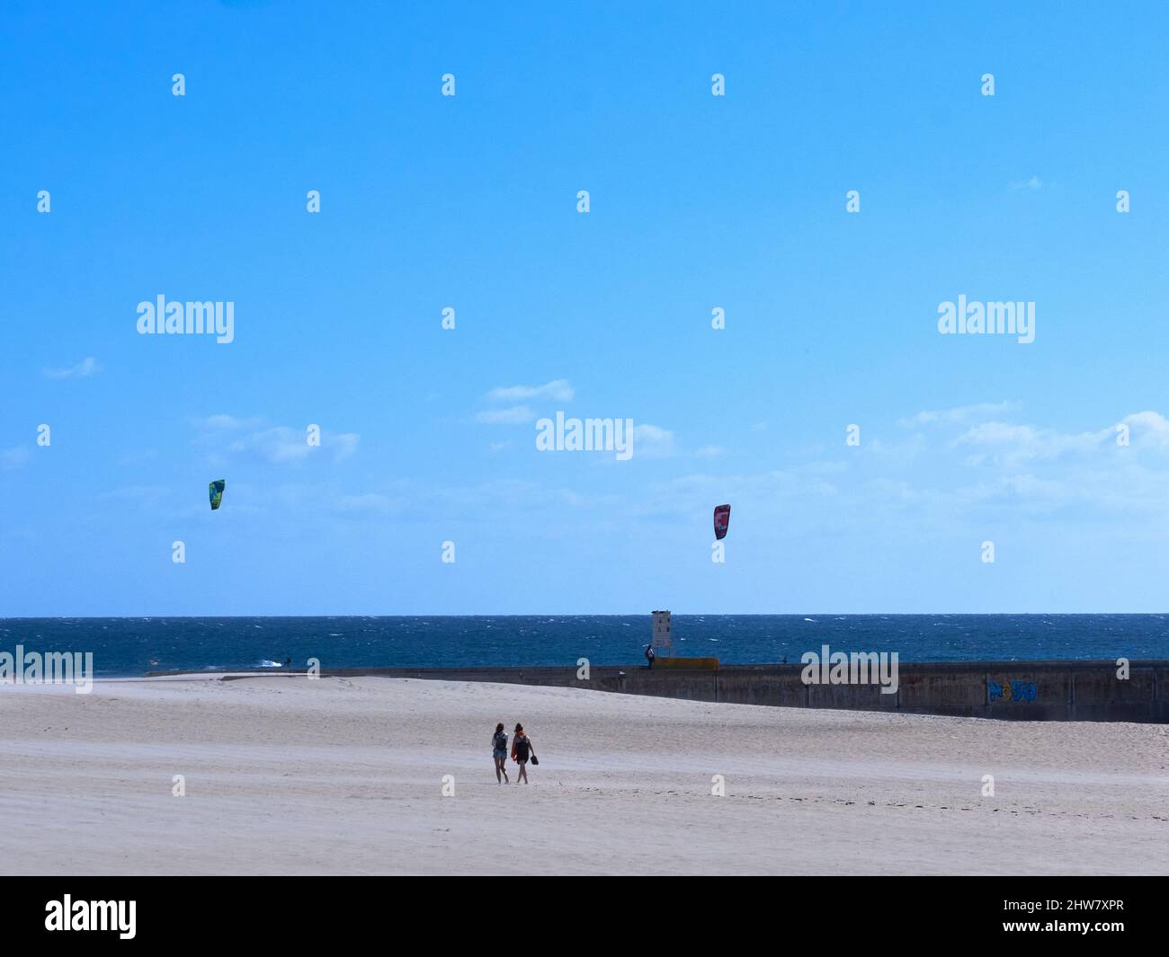 Zwei Personen, die am Strand von Barra gegen die Kitesurfer in blauer Meereslandschaft in Aveiro, Portugal, wandern Stockfoto