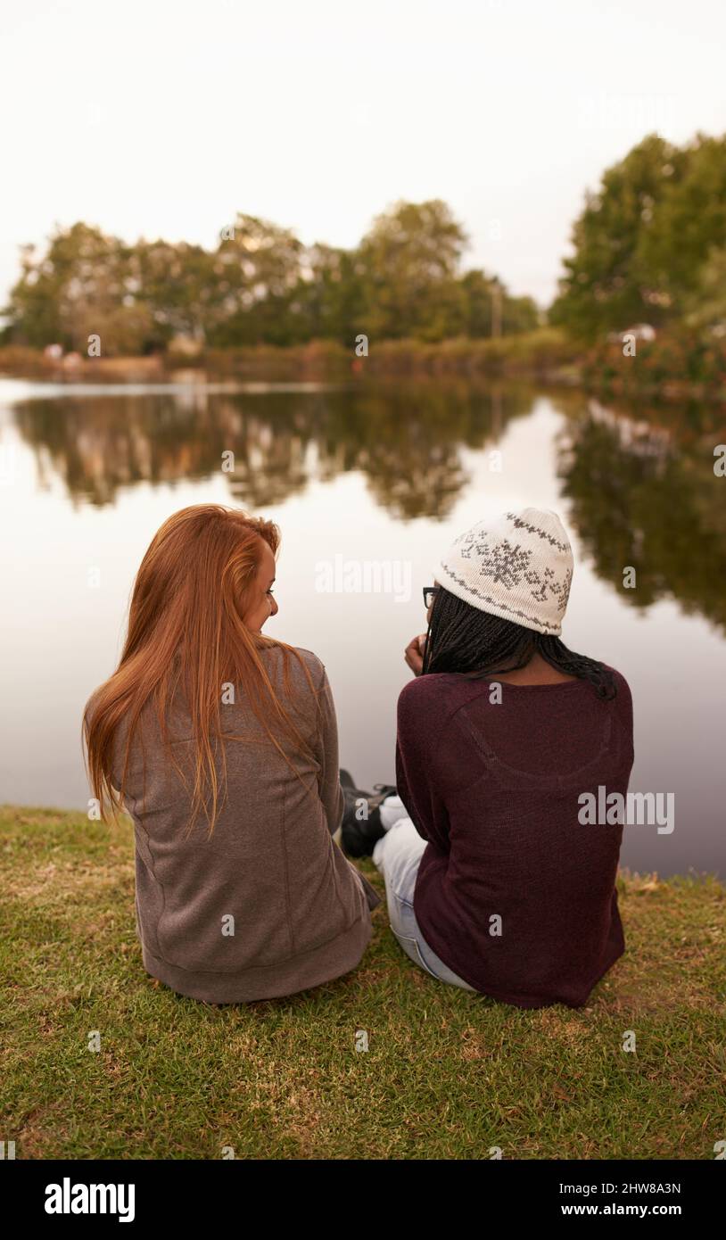 Gute Zeit mit meinem Partner in der Kriminalität. Rückansicht von zwei Mädchen im Teenageralter, die neben einem See sitzen und sich unterhalten. Stockfoto