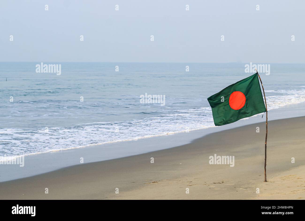 Flagge Bangladeschs am Strand bei Sonnenaufgang oder Sonnenuntergang. Bangladeschische Nationalflagge am Sommerstrand mit Sand und Welle. Sommerhintergrund. Stockfoto