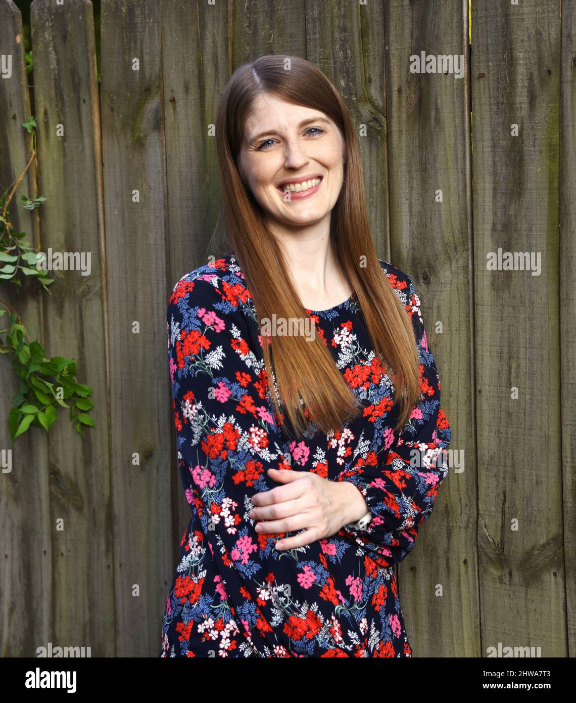 Die junge Frau kann den Tag mit ihrem großen Lächeln erhellen. Sie trägt eine bedruckte Bluse und hat lange braune Haare. Sie steht neben einem rustikalen Holzzaun Stockfoto