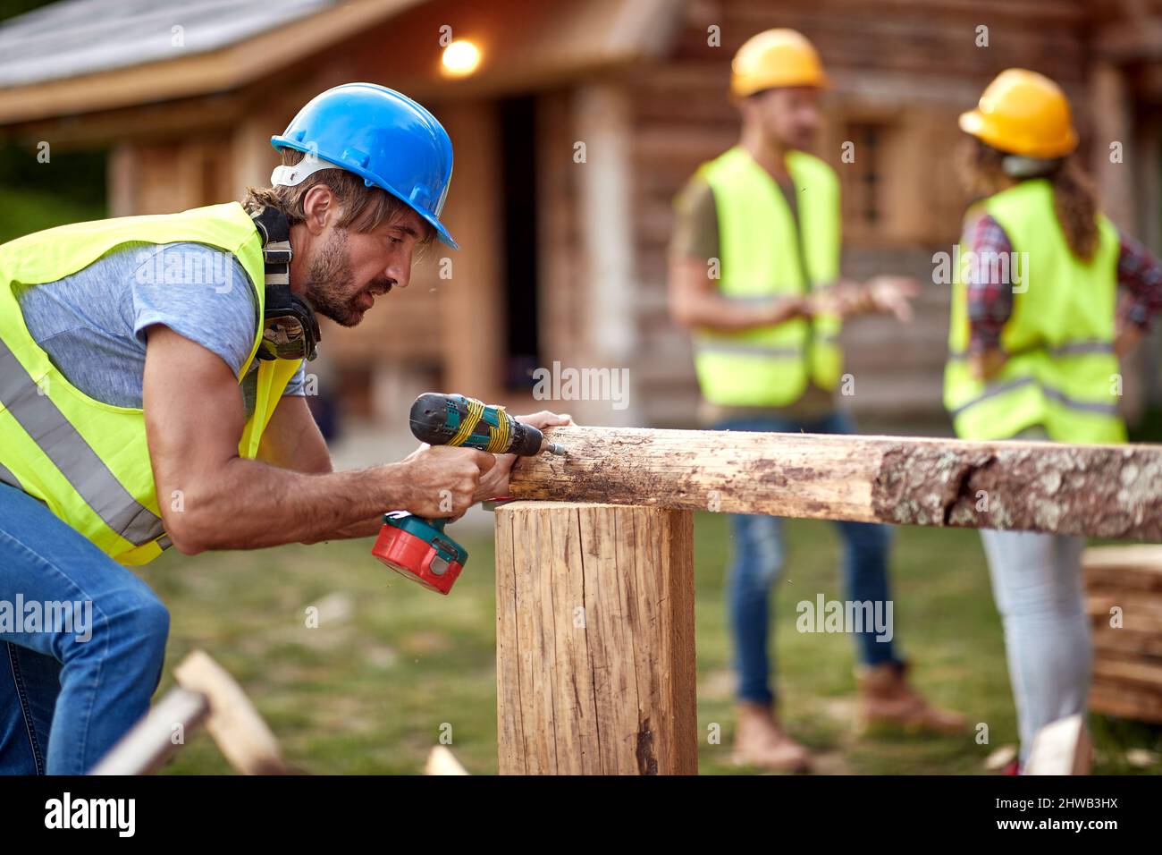 Junger Bauarbeiter, der mit Bohrmaschine auf Holzkonstruktion arbeitet. Andere Mitarbeiter, die im Hintergrund ein Gespräch führen. Holzskelett Stockfoto