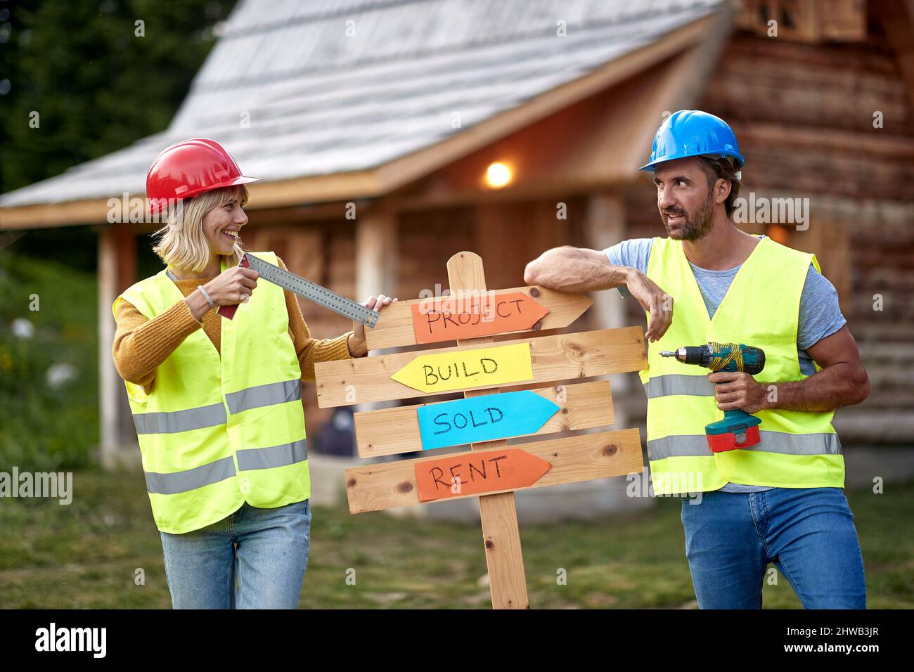 Fröhliches, kraftvolles junges Baupaar mit Holzbrett. Schmunzelend, fröhlich, vor einem Holzhaus. Teamarbeit. Holzskelettframing Building. Stockfoto