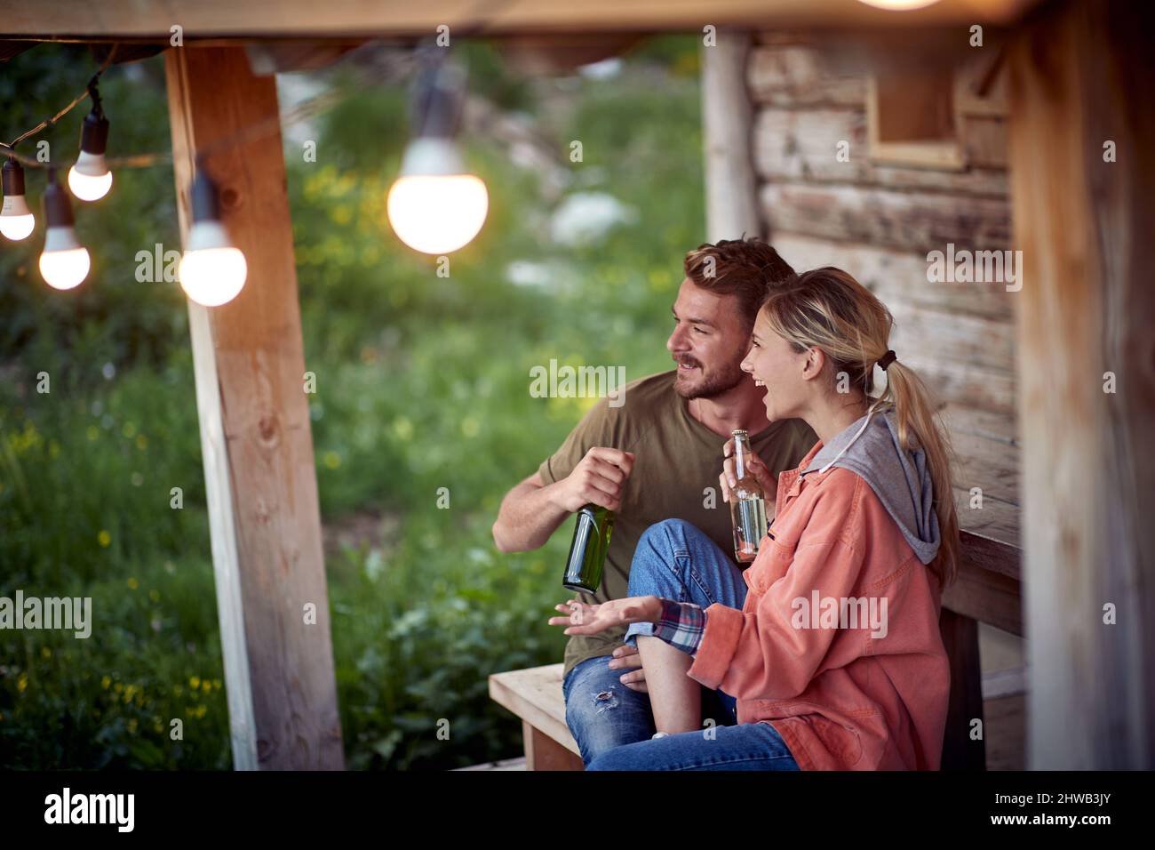 Ein junges, fröhliches Paar, das vor einem Holzhäuschen auf der Terrasse ein Gespräch führt und Bier trinkt. Sommer Gartenfest und Spaß. Freund Stockfoto