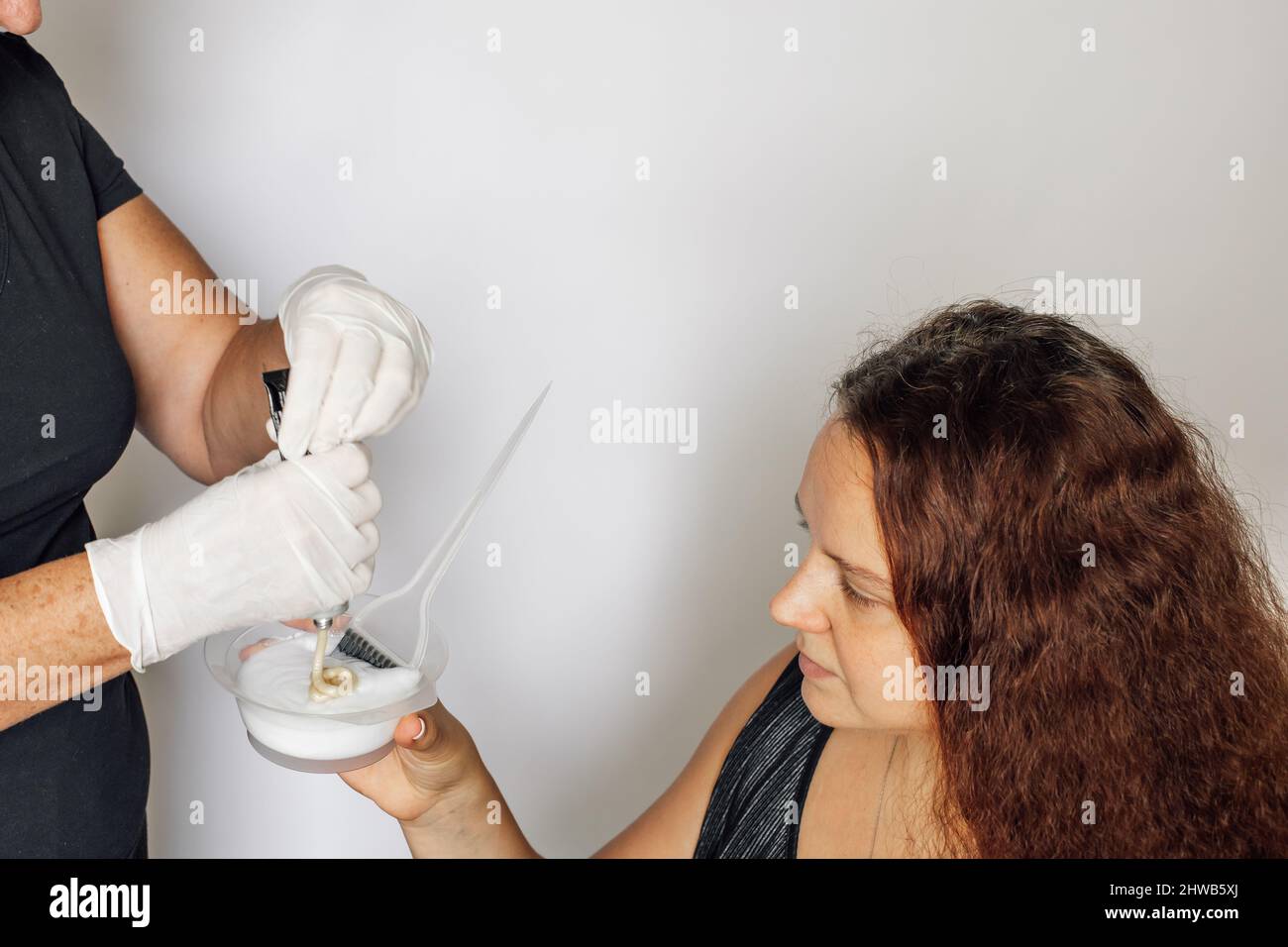 Weibliche Hände in Latexhandschuhen, die den Inhalt des Haarfärberohrs in die Schüssel quetschen, in der Hand einer rothaarigen Frau, die die Farbsubstanz auf Weiß ansieht Stockfoto