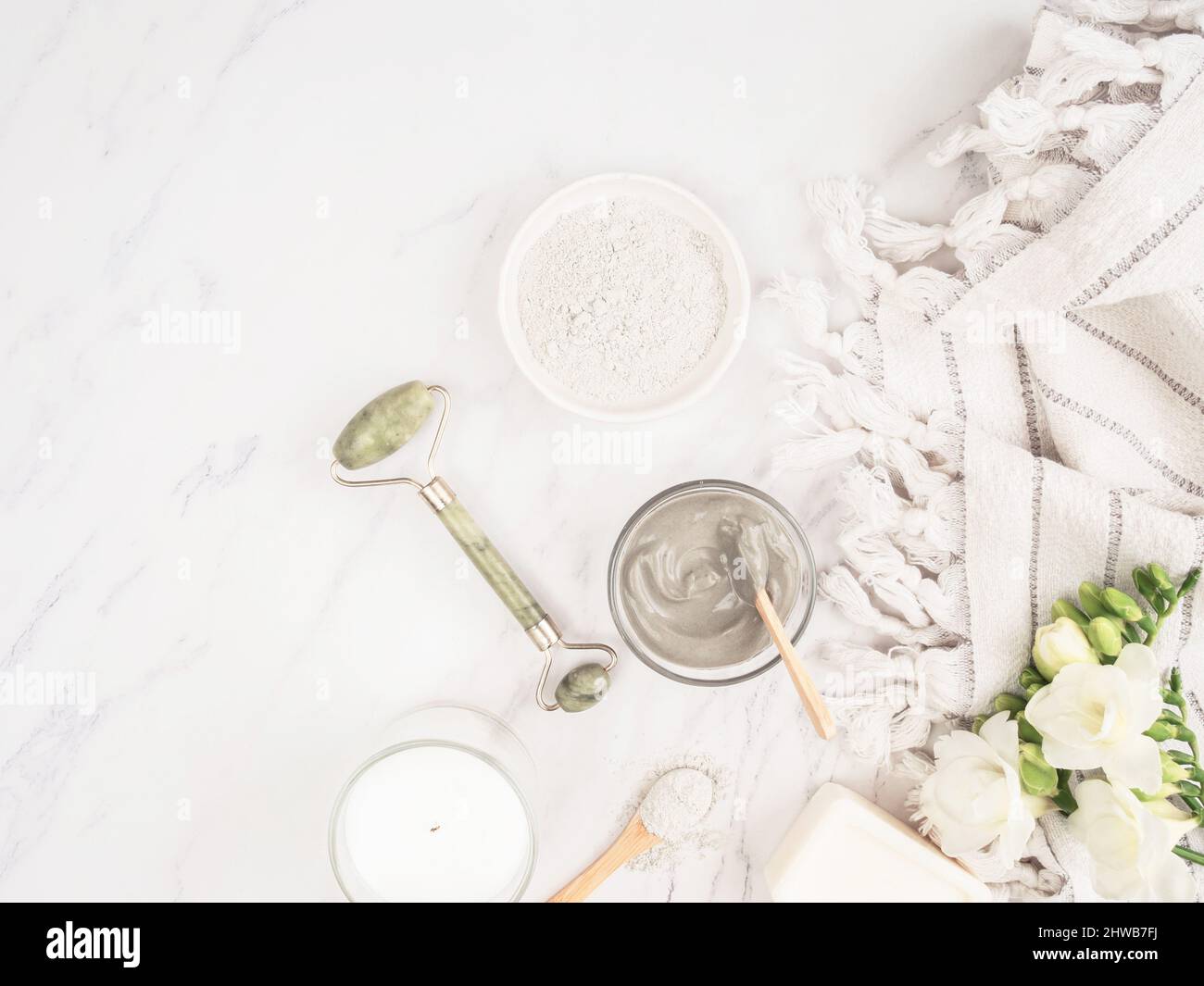 Hausgemachtes Hautpflegekonzept. Bio-blauer Ton für Beauty-Gesichtsmaske, Kristall-Massagegerät, Kerze, Baumwolltuch und weiße Blumen auf Marmor-Hintergrund. T Stockfoto