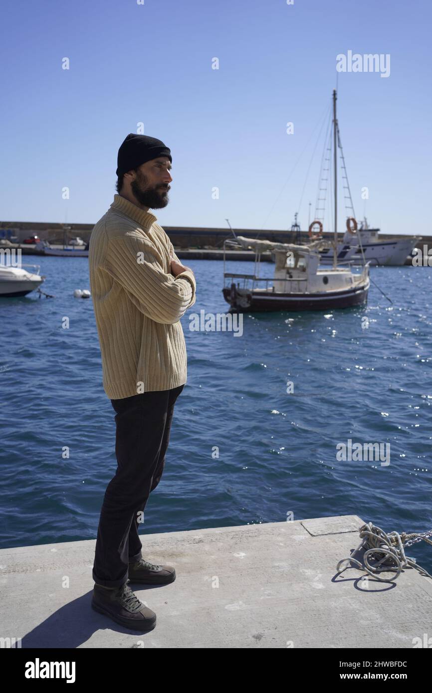 Auf einem Schiff in einem Seehafen steht ein großer und robuster junger Seemann mit einem Bart in einer Wollmütze und einem hochhalsigen Pullover Stockfoto