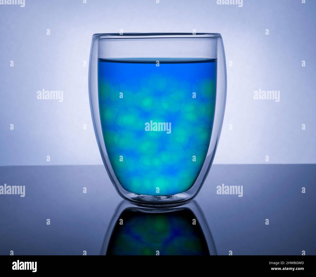 Doppelwandiger isolierter Glasbecher, gefüllt mit blauer und grüner Flüssigkeit und rundem Eis.heller reflektierender Hintergrund Stockfoto