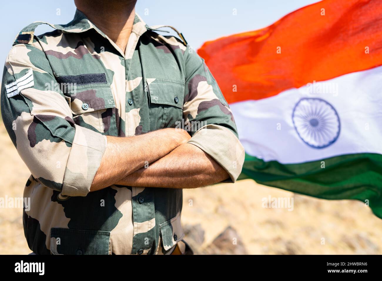 Indischer Soldat, der mit gekreuzten Armen vor der winkenden indischen Flagge steht - Konzept eines selbstbewussten, furchtlosen und stolzen Soldaten Stockfoto