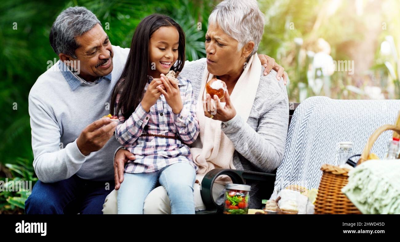 Wir haben immer gesunde Leckereien für sie. Aufnahme eines fröhlichen kleinen Mädchens, das mit ihren Großeltern im Park auf einer Bank sitzt. Stockfoto