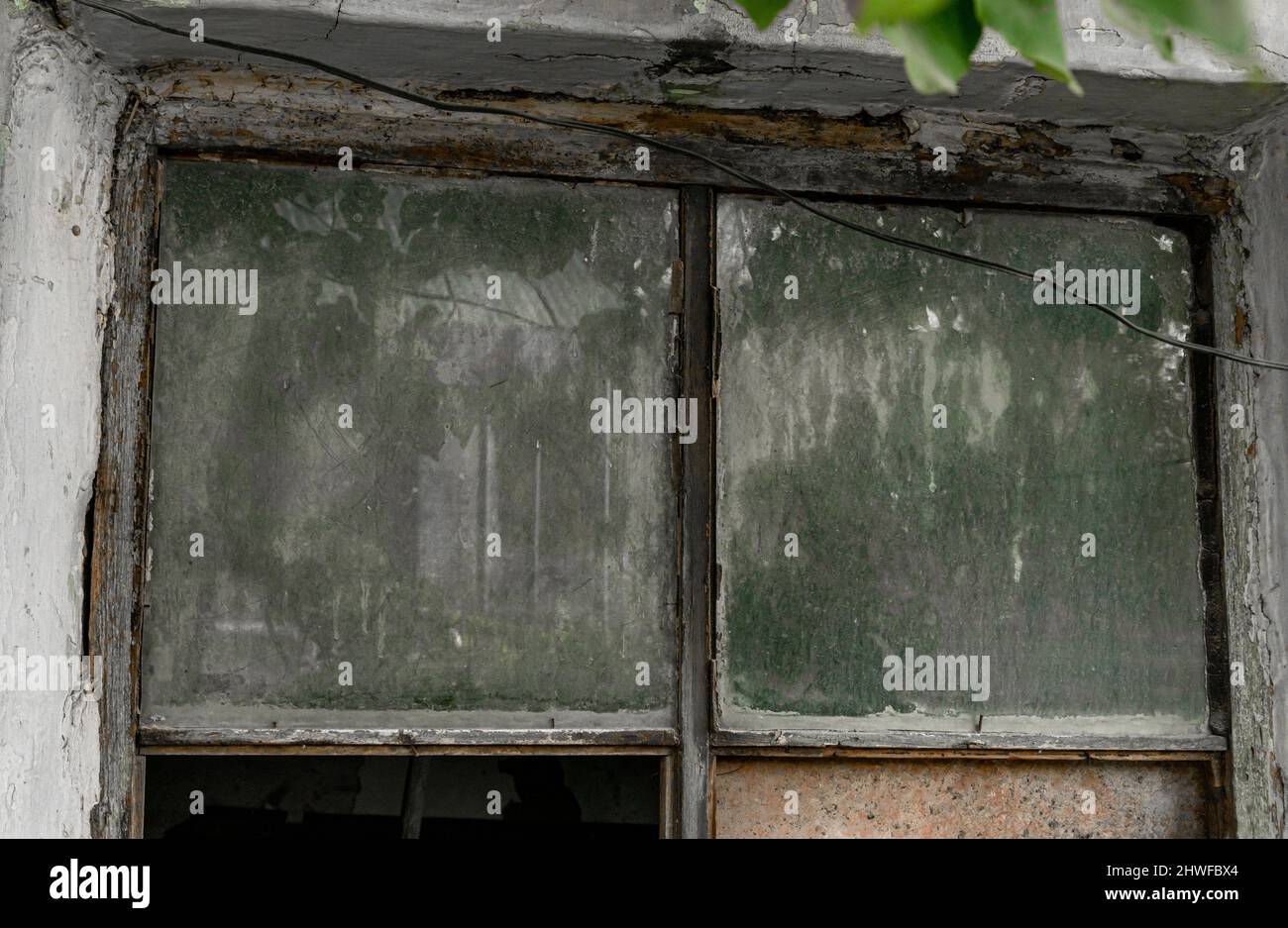 Kleines, gerahmtes, staubiges Fenster in einer Wand mit zerlegter Verputzung, die Schichten aus сement und Backstein zeigt. Schmutziges ungewaschene Fenster. Stockfoto