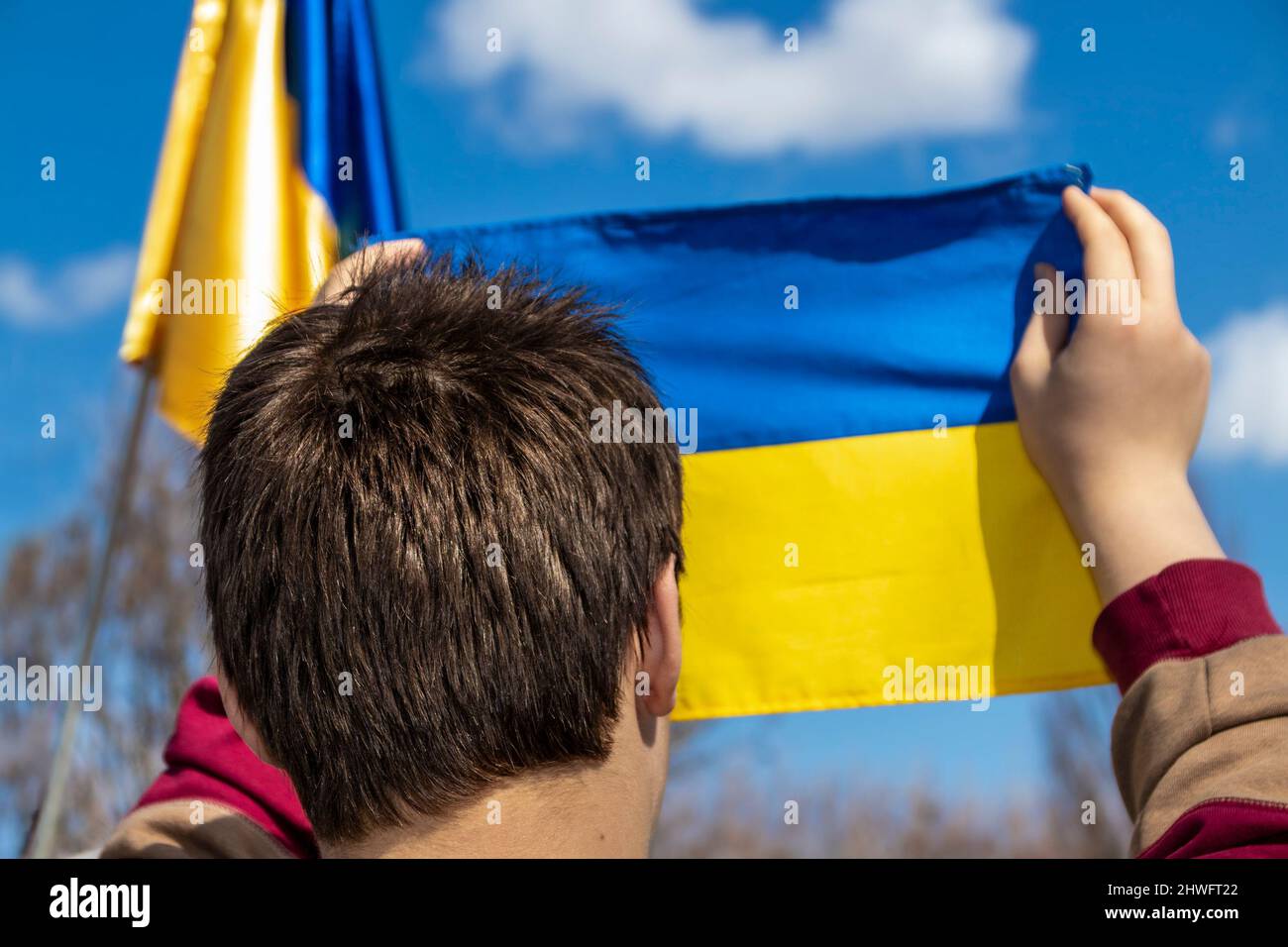 Leiter eines jungen Mannes, der bei einer Kundgebung im Freien eine kleine ukrainische Flagge hochhält, mit einer weiteren verschwommenen Flagge im Hintergrund – selektiver Fokus Stockfoto