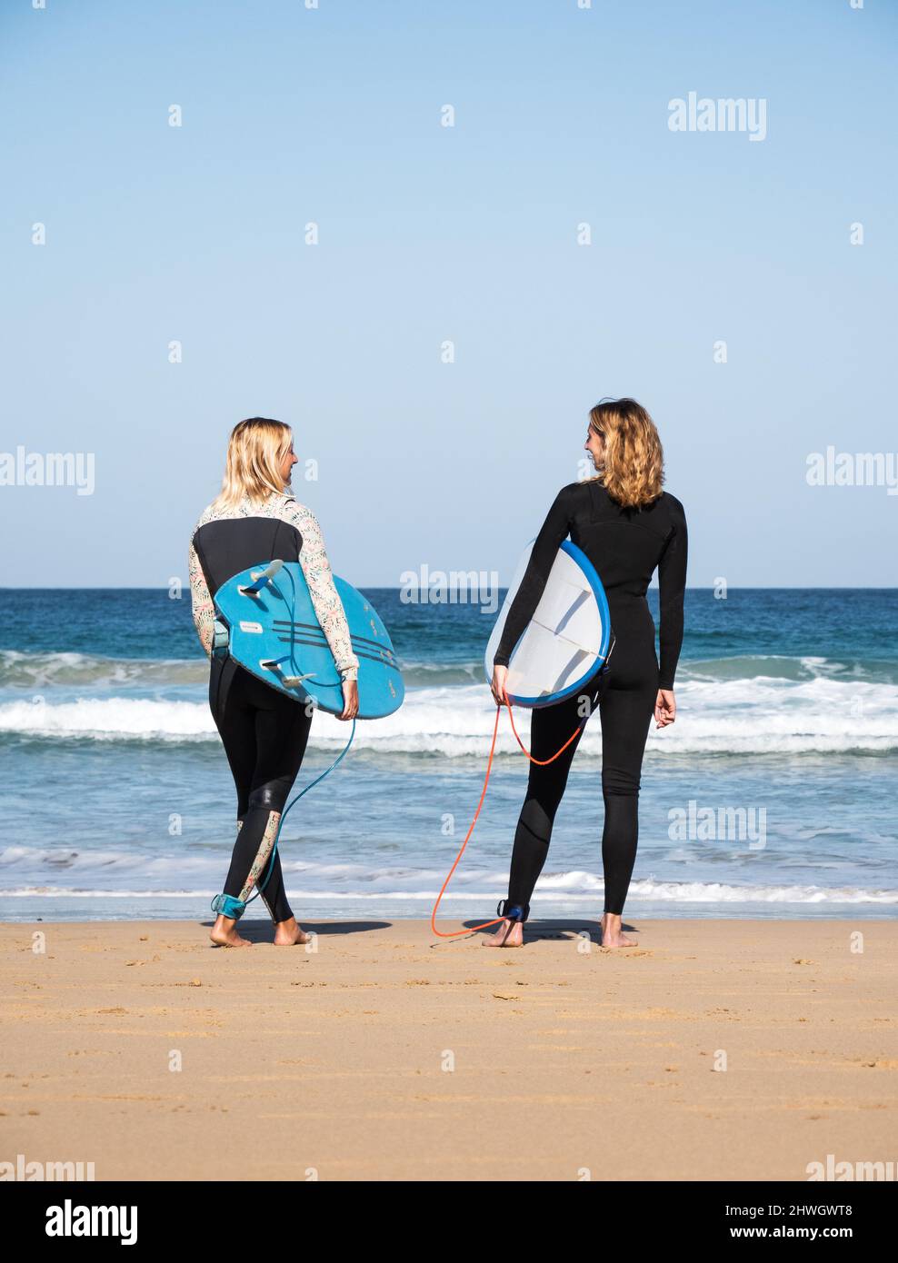 2 kaukasische Surferinnen stehen am Strand und beobachten die Wellen. Sie tragen Winter-Neoprenanzüge und halten Surfbretter. Stockfoto