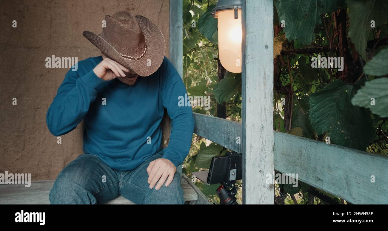 Cowboy-Mann sieht Comedy auf seinem Handy und lacht auf der Veranda des Hauses sitzen Stockfoto