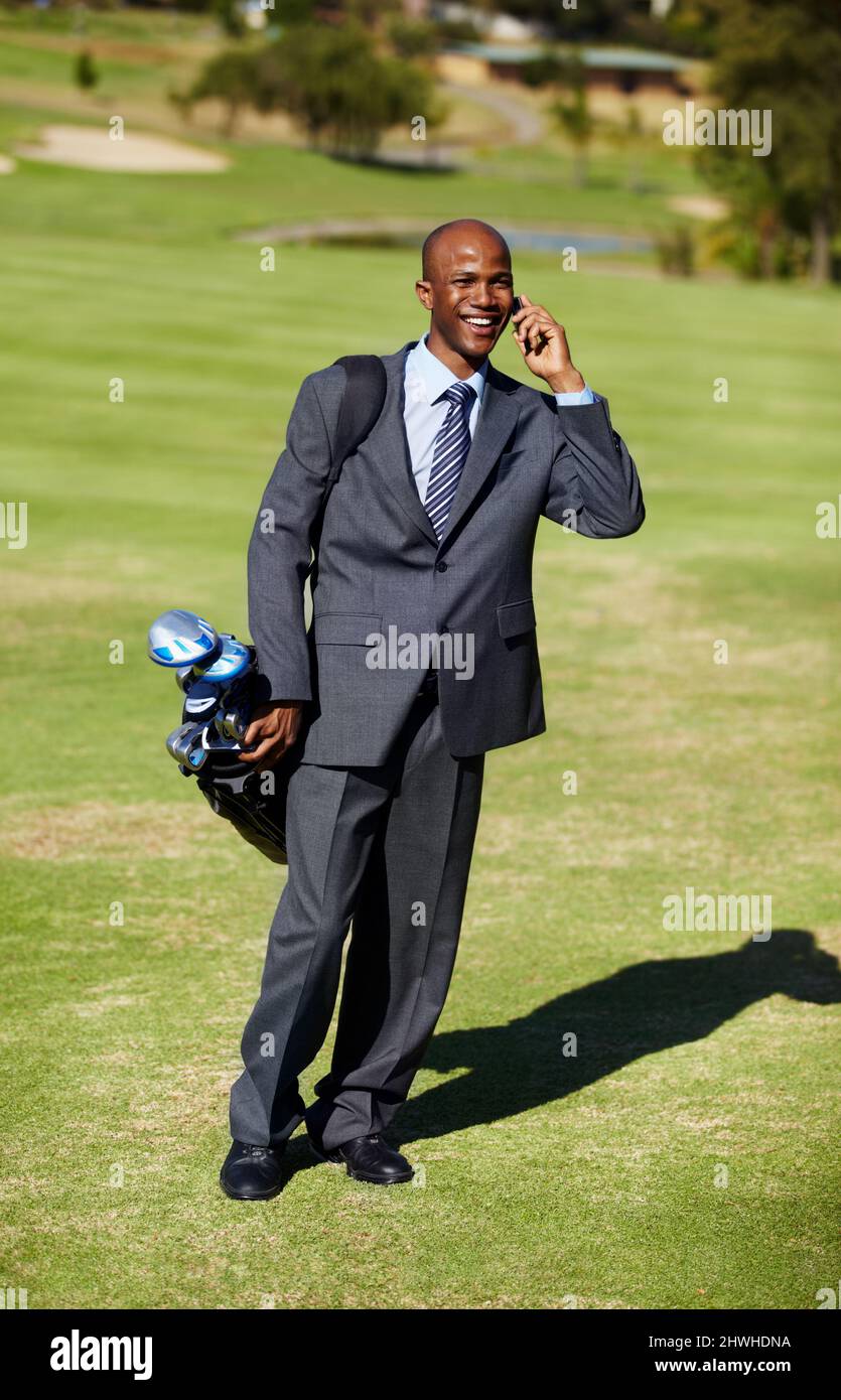 Geschäfte auf dem Kurs machen. Aufnahme eines afrikaners, der einen Anzug trägt und am Telefon spricht, während er eine Golftasche auf einem Golfplatz trägt. Stockfoto